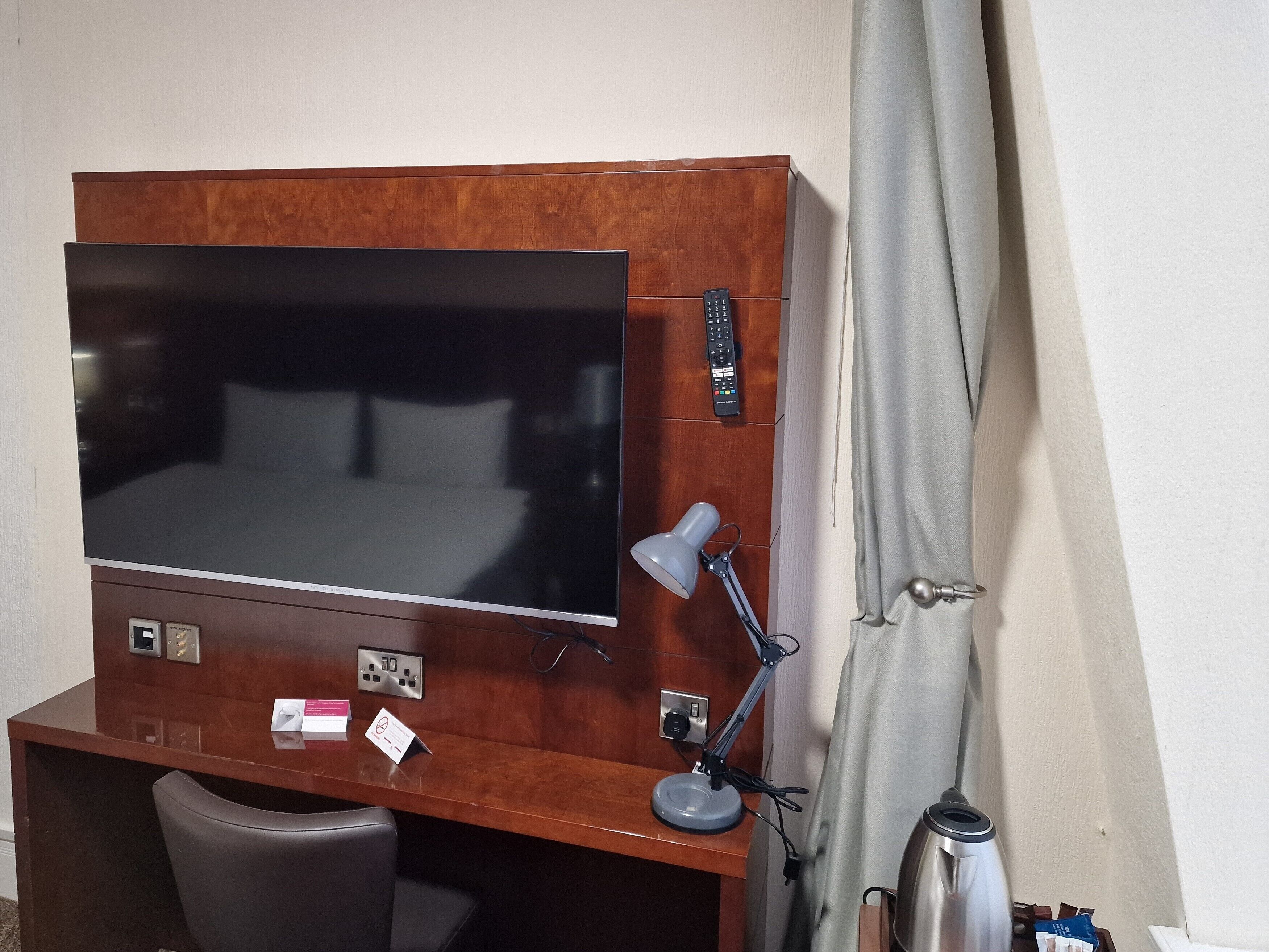 A hotel room with a wooden desk, a flat-screen TV, a desk lamp, a chair, and a coffee maker.