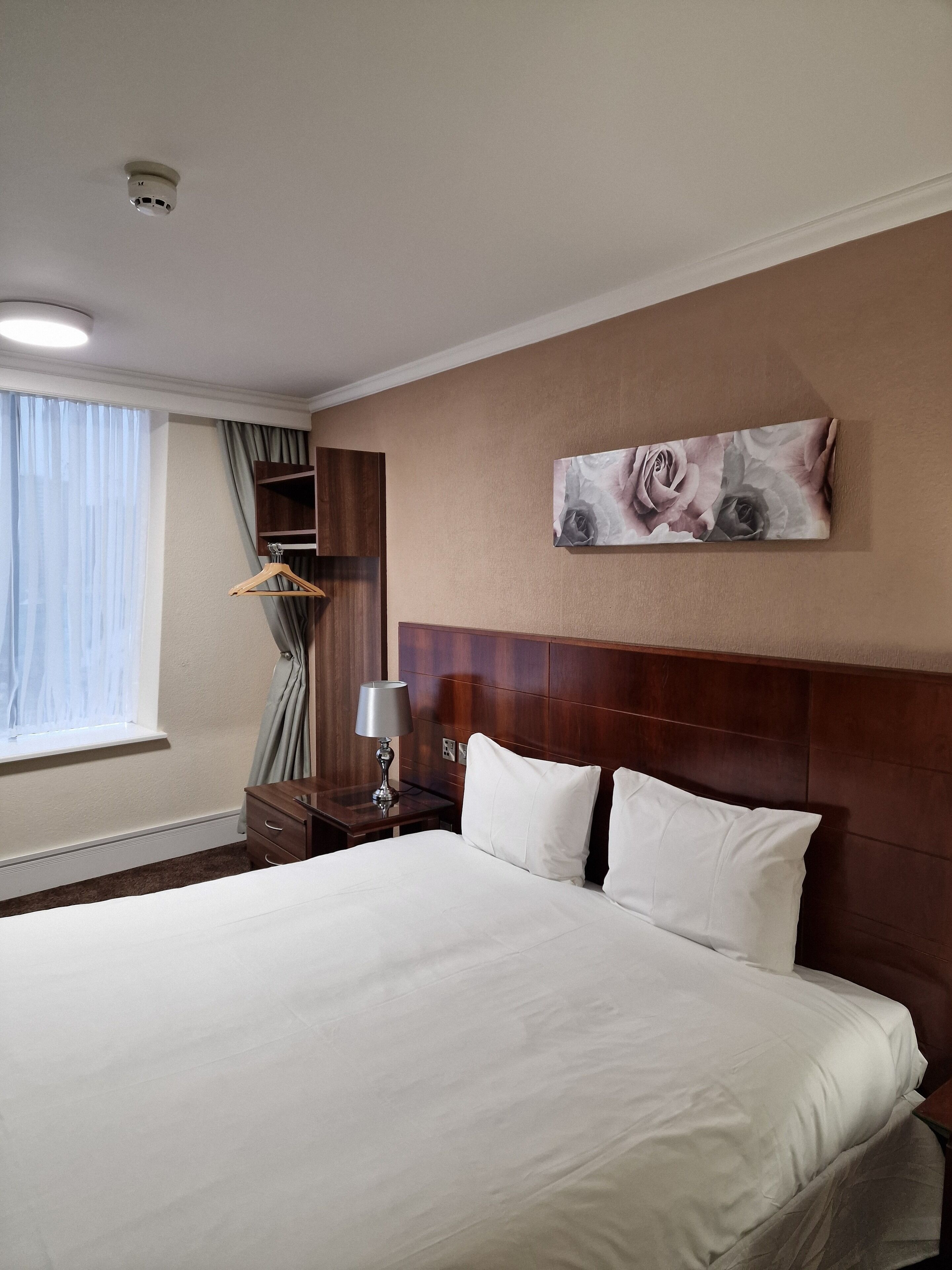 A hotel room with a large bed, a wooden headboard, a nightstand with a lamp, a wardrobe, and a window with curtains.