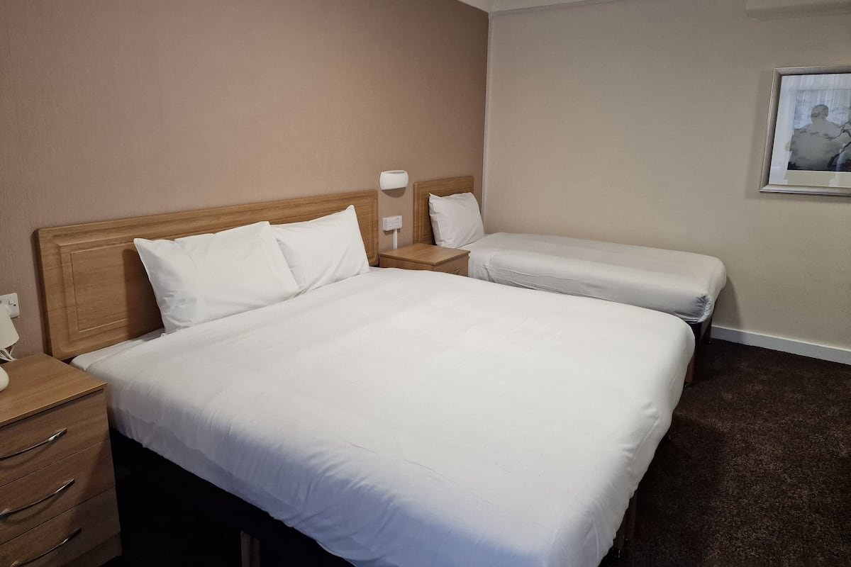 Habitación de hotel con dos camas individuales, cabeceros de madera, una mesita de noche con lámpara y un cuadro en la pared.