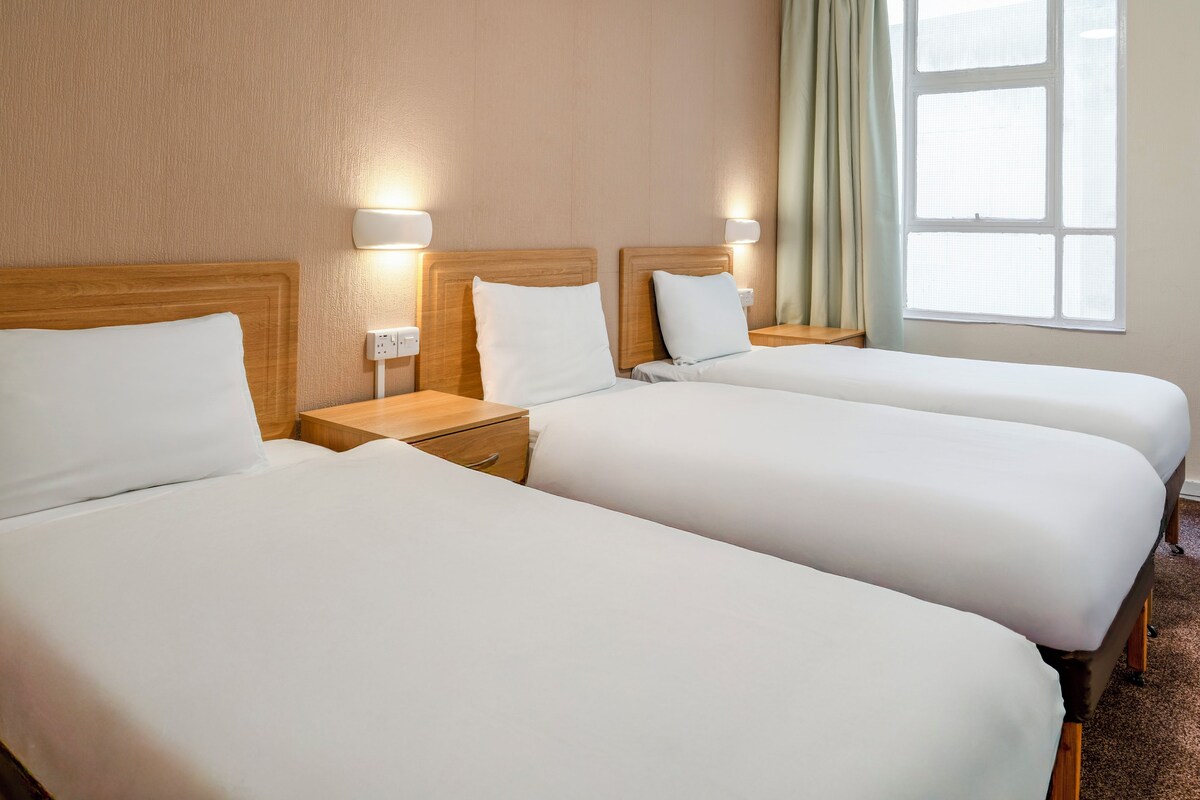 Habitación de hotel con dos camas individuales, cabeceros de madera, mesitas de noche y una ventana con cortinas.