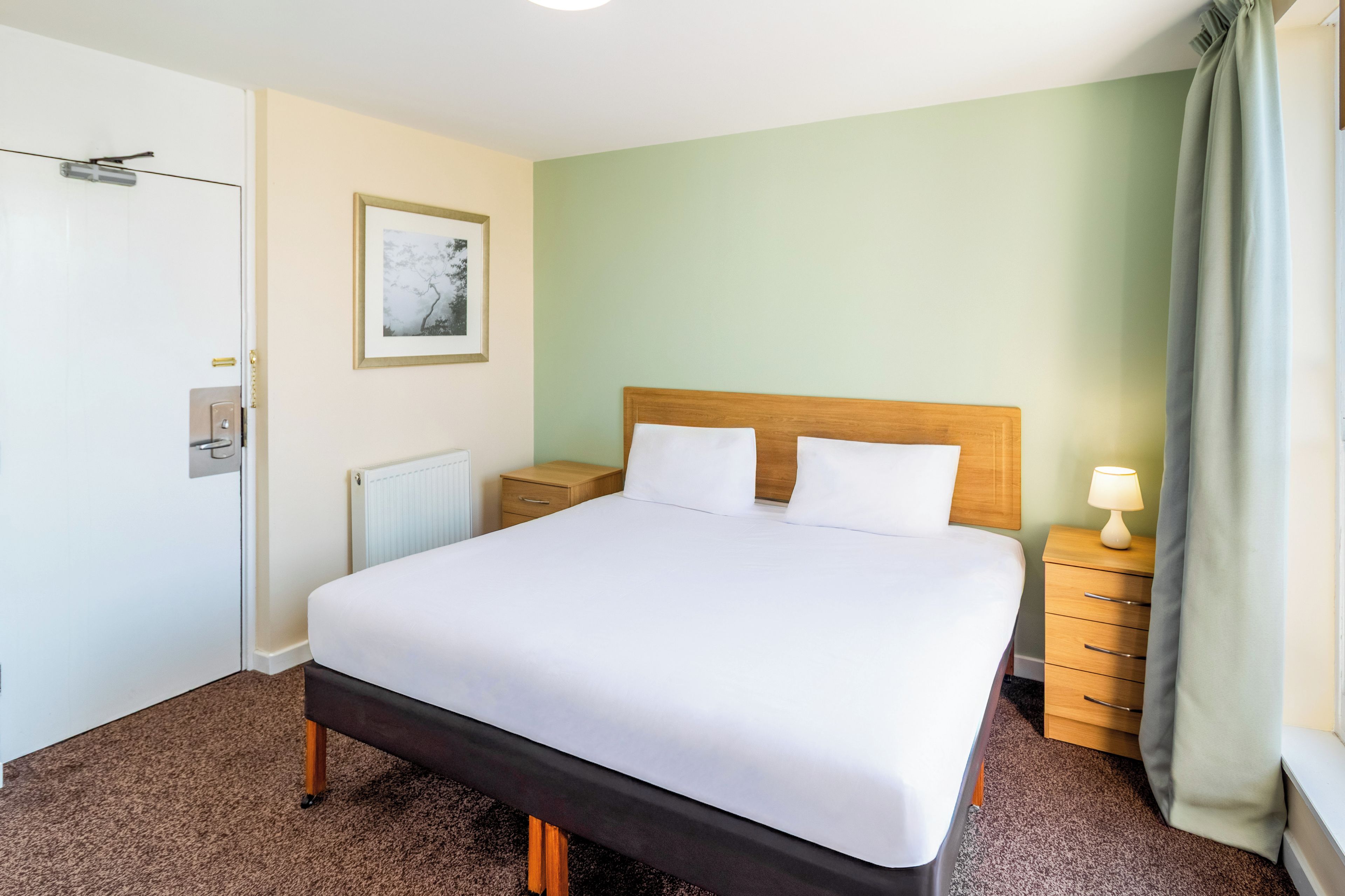 A hotel room with a bed, a bedside table with a lamp, a radiator, a framed picture on the wall, and a door.