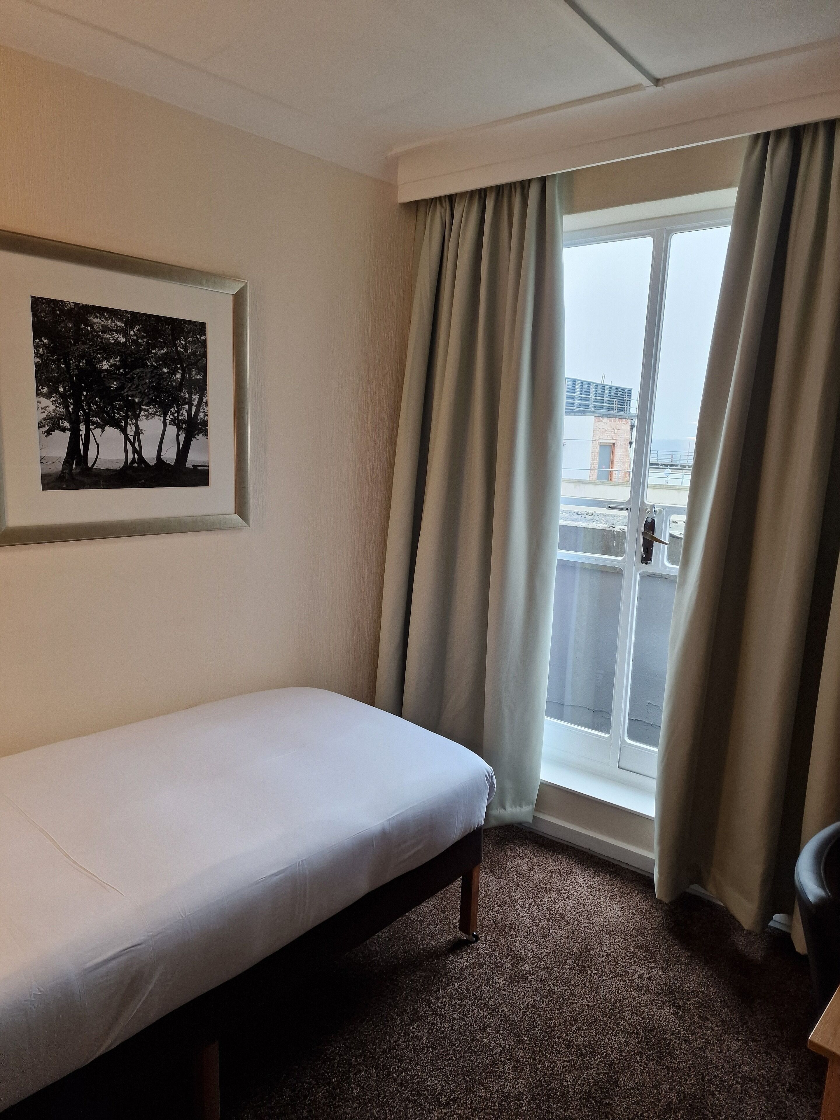 Hotellihuone, jossa on yksi sänky, seinällä kehystetty taulu ja ikkunasta avautuva näkymä kaupunkiin.
