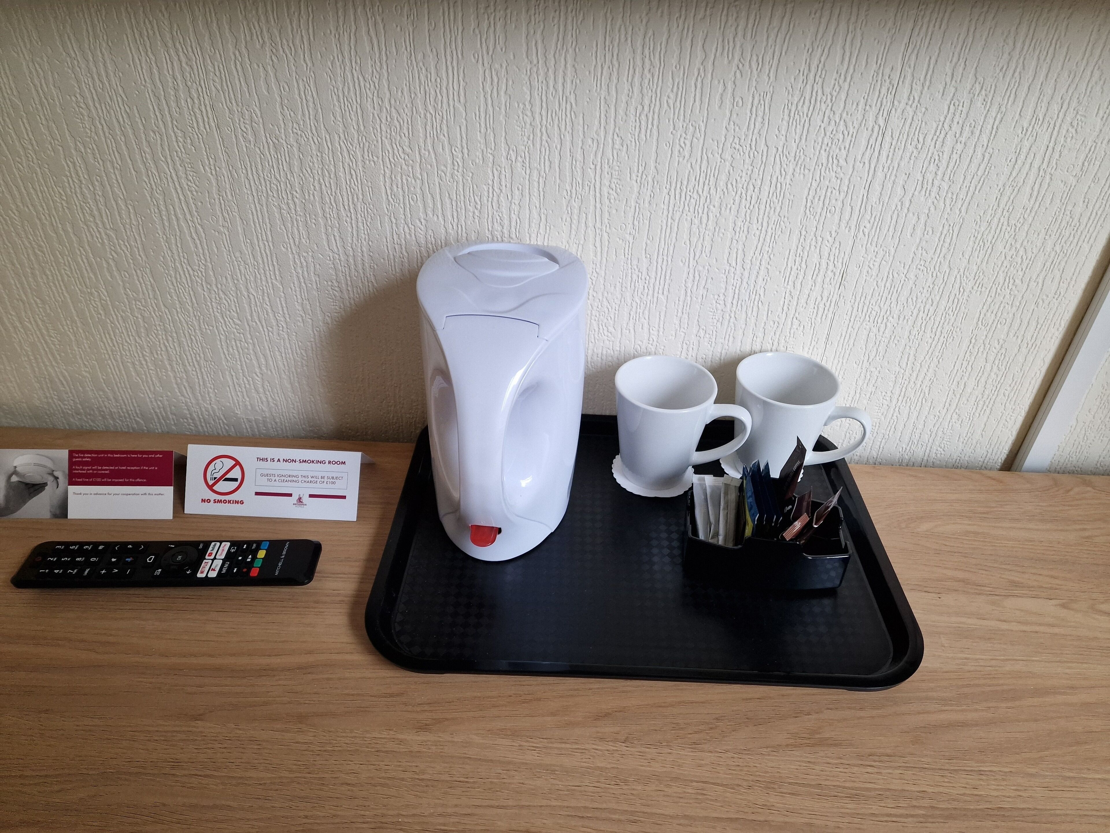 A non-smoking room sign, a white electric kettle, two white mugs, a remote control, and a tray with pens and a notebook.