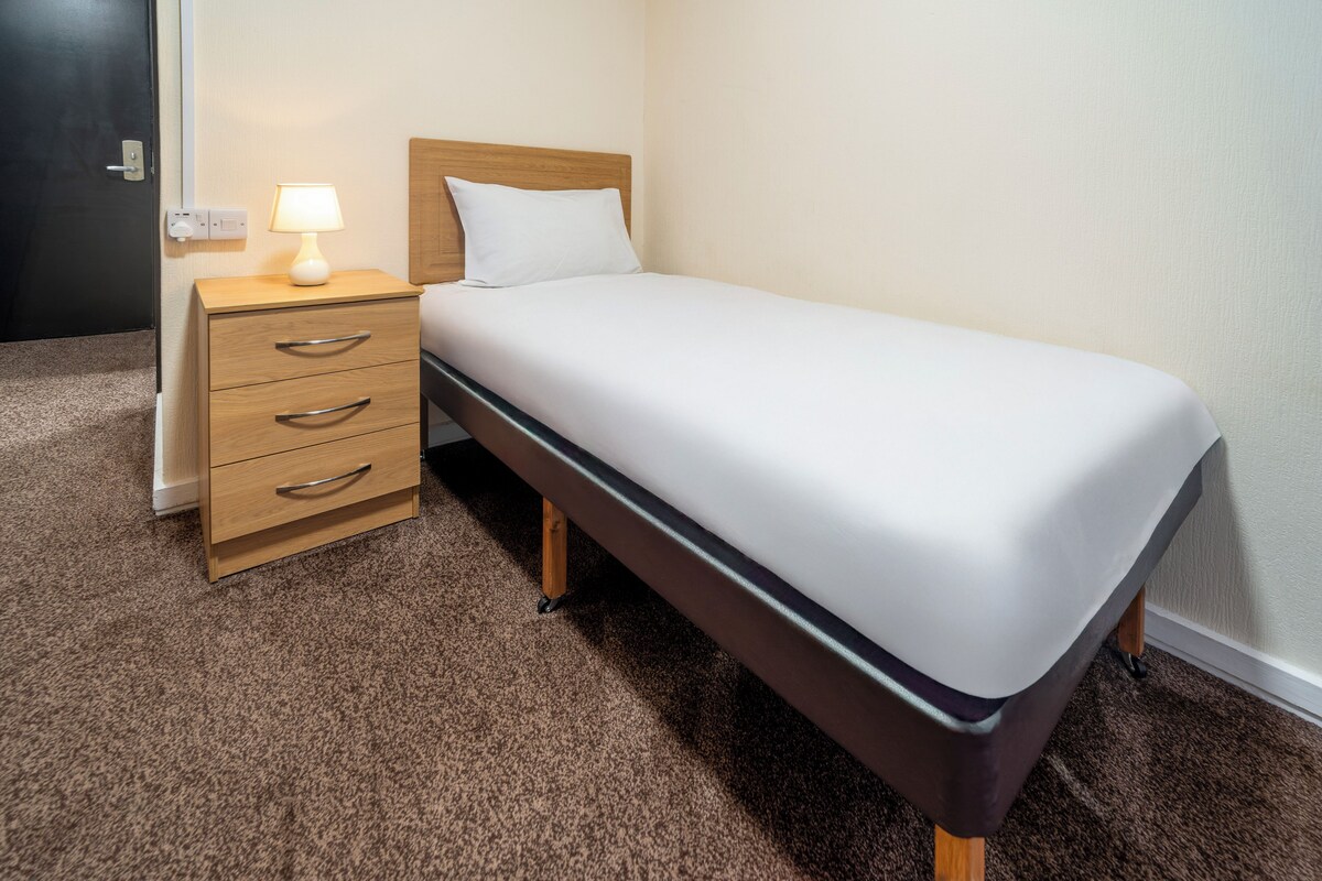 Una cama individual con cabecera de madera, una mesita de noche con cajones y una lámpara.