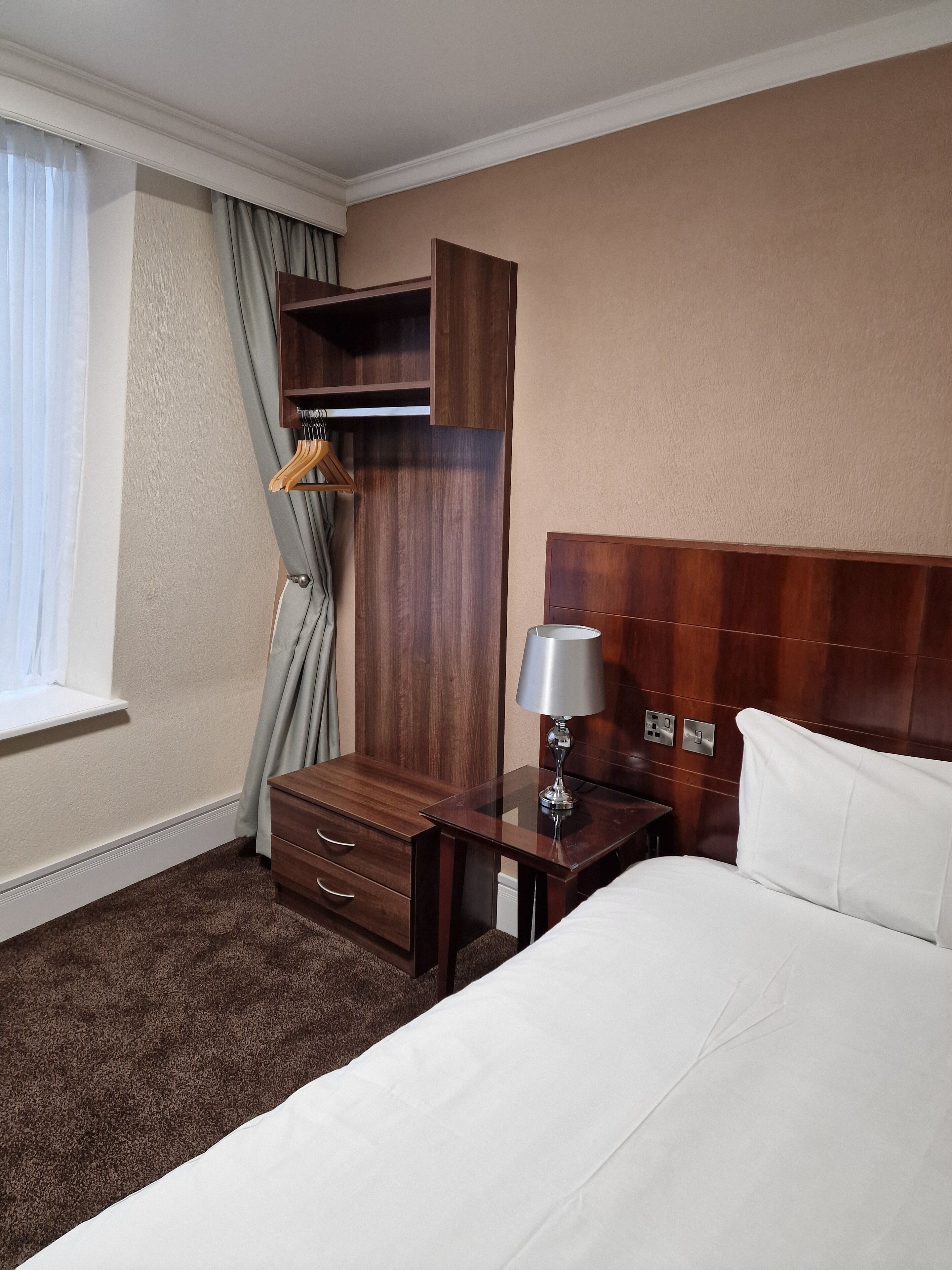 A hotel room with a bed, a nightstand, a wooden wardrobe, a lamp, and a window with curtains.