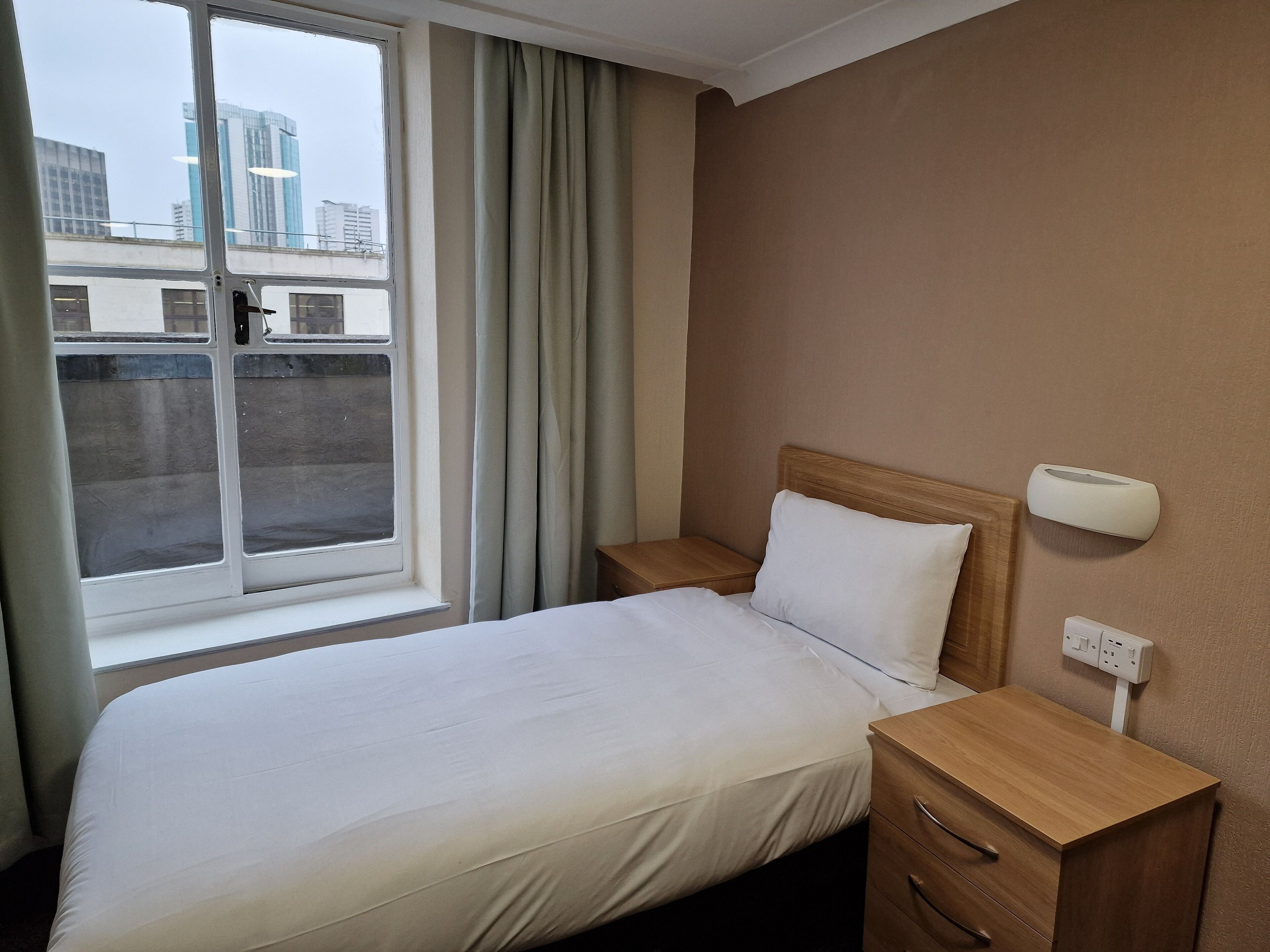 A hotel room with a bed, wooden nightstands, a bedside table, a wall-mounted light, and a window with curtains.