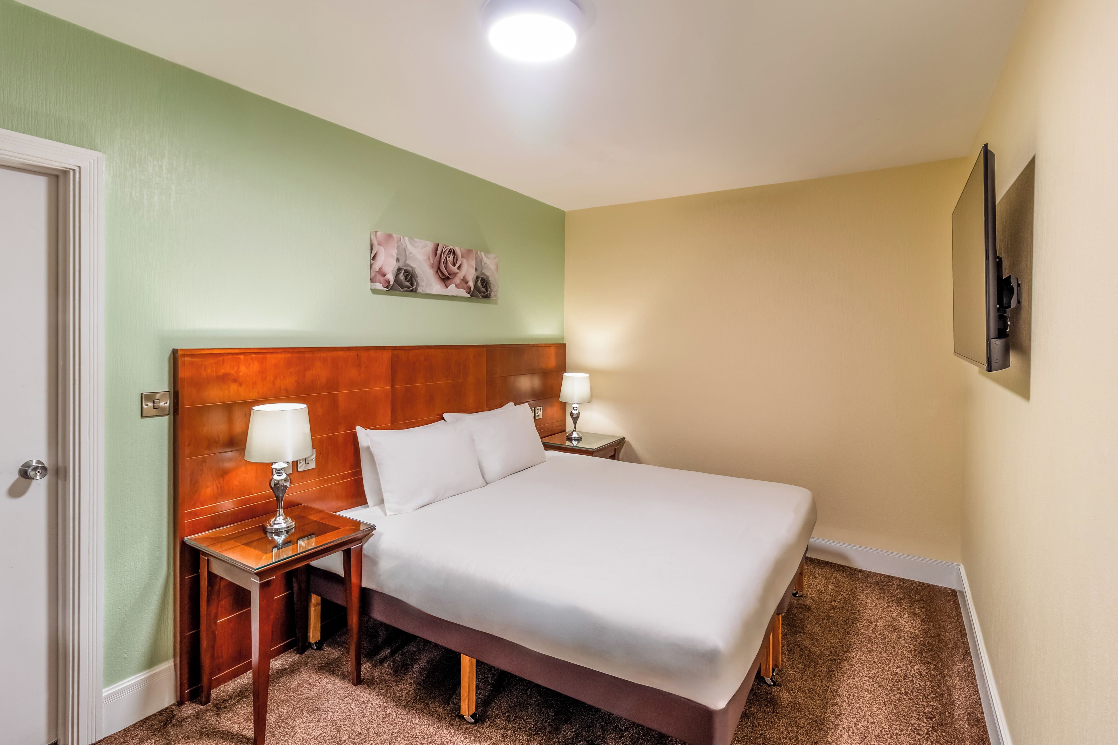 Una habitación de hotel con cama, dos mesitas de noche con lámparas, un televisor en la pared y una puerta.