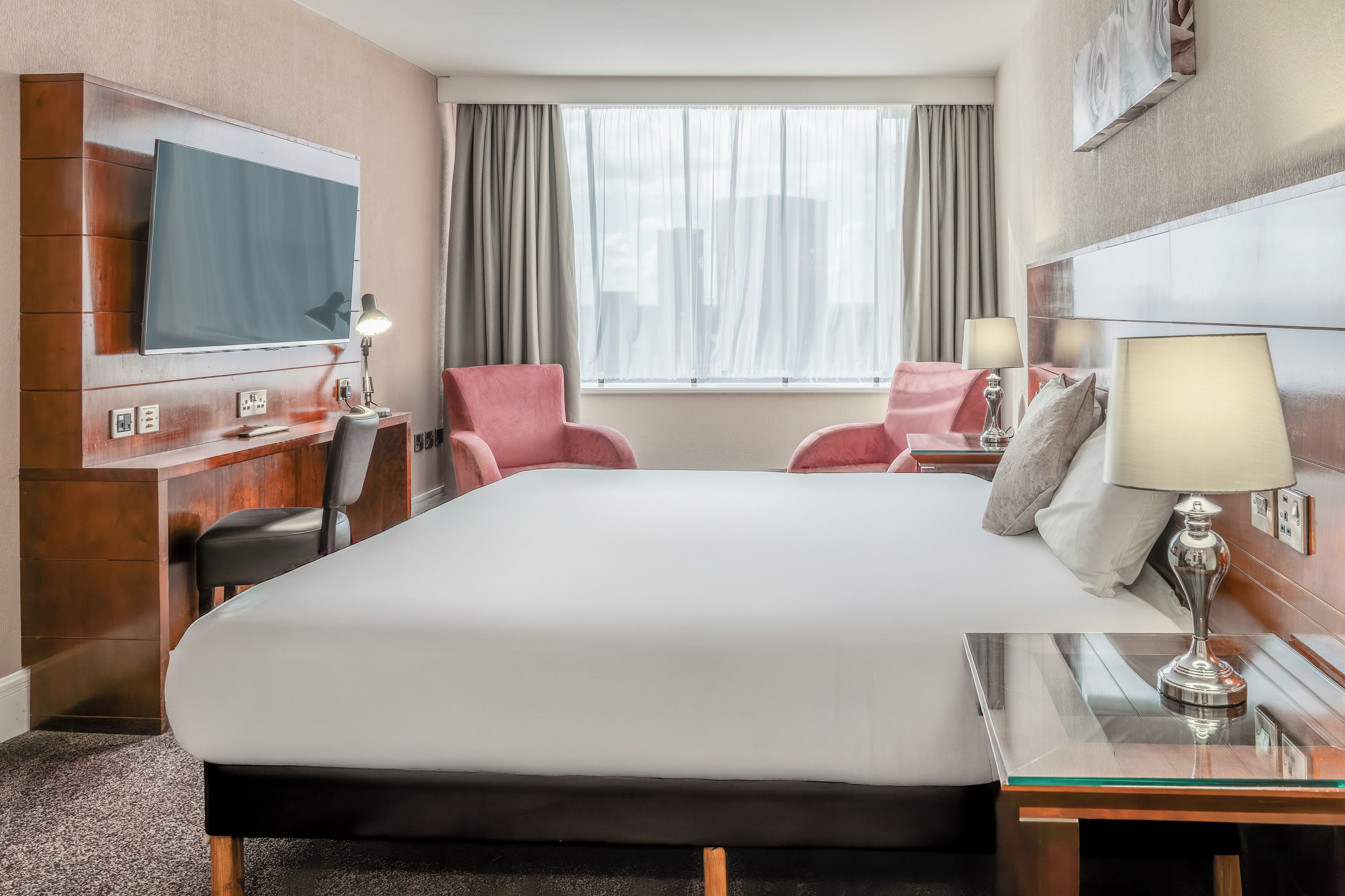 A hotel room with a large bed, a desk with a lamp, a chair, a TV, and a window with curtains.
