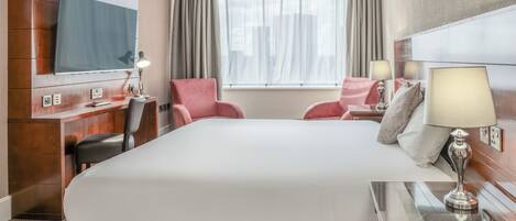 A hotel room with a large bed, a desk with a lamp, a chair, a TV, and a window with curtains.