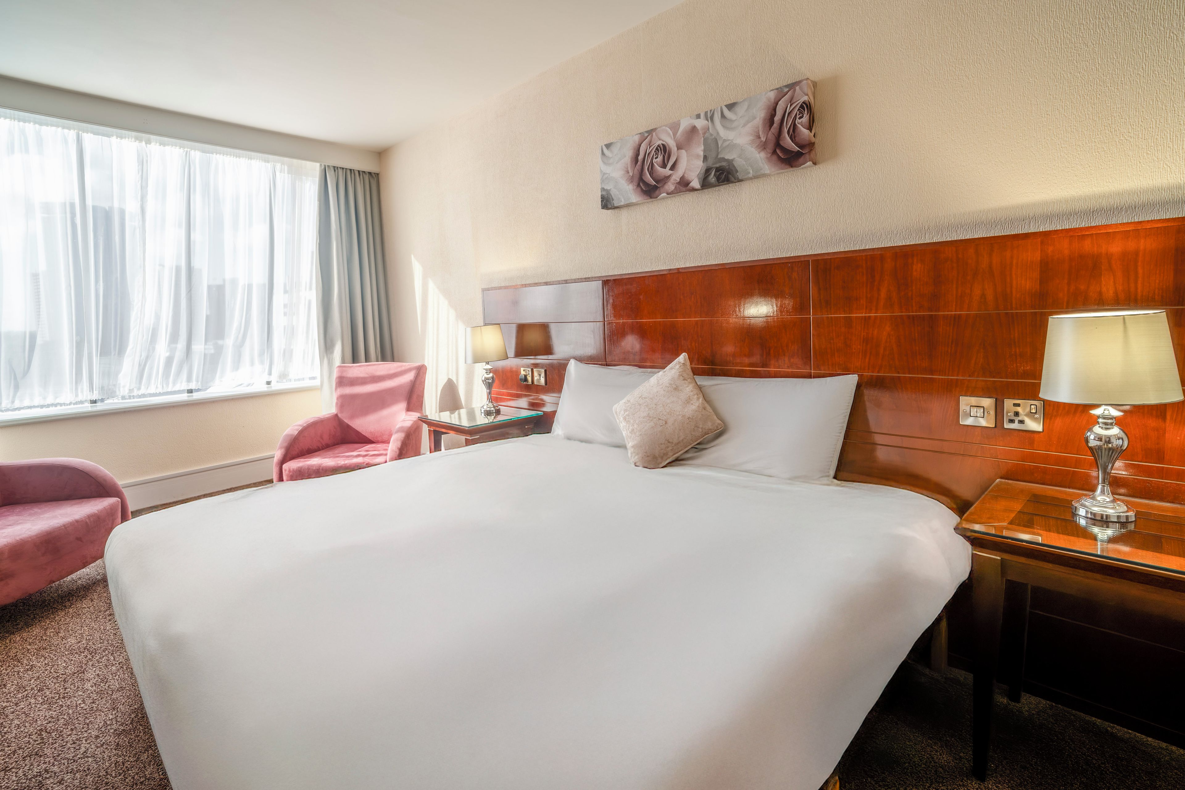 A hotel room with a large bed, a wooden headboard, a nightstand with a lamp, a chair, and a window with curtains.