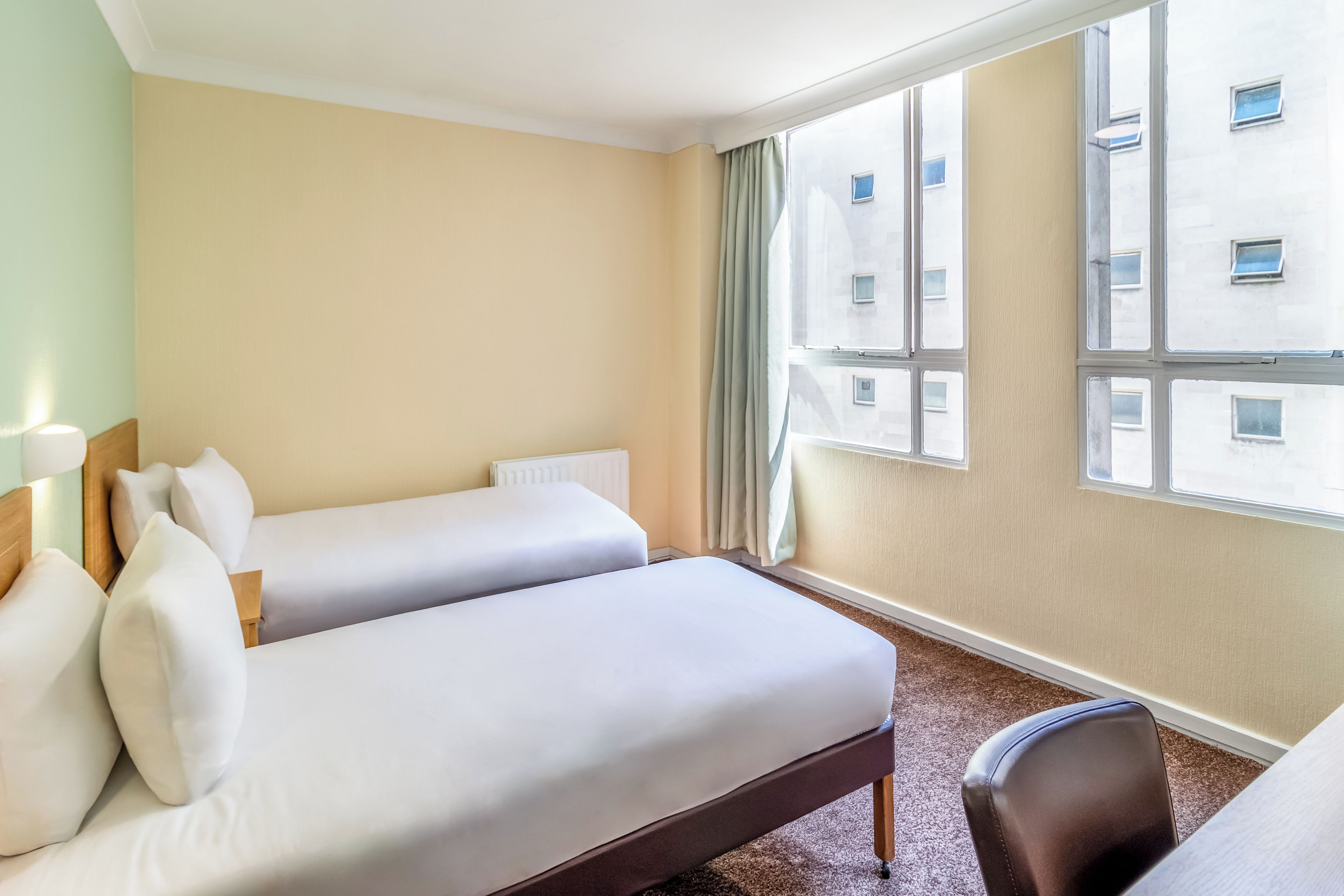 A hotel room with two single beds, a wooden headboard, a window with curtains, and a view of a building.