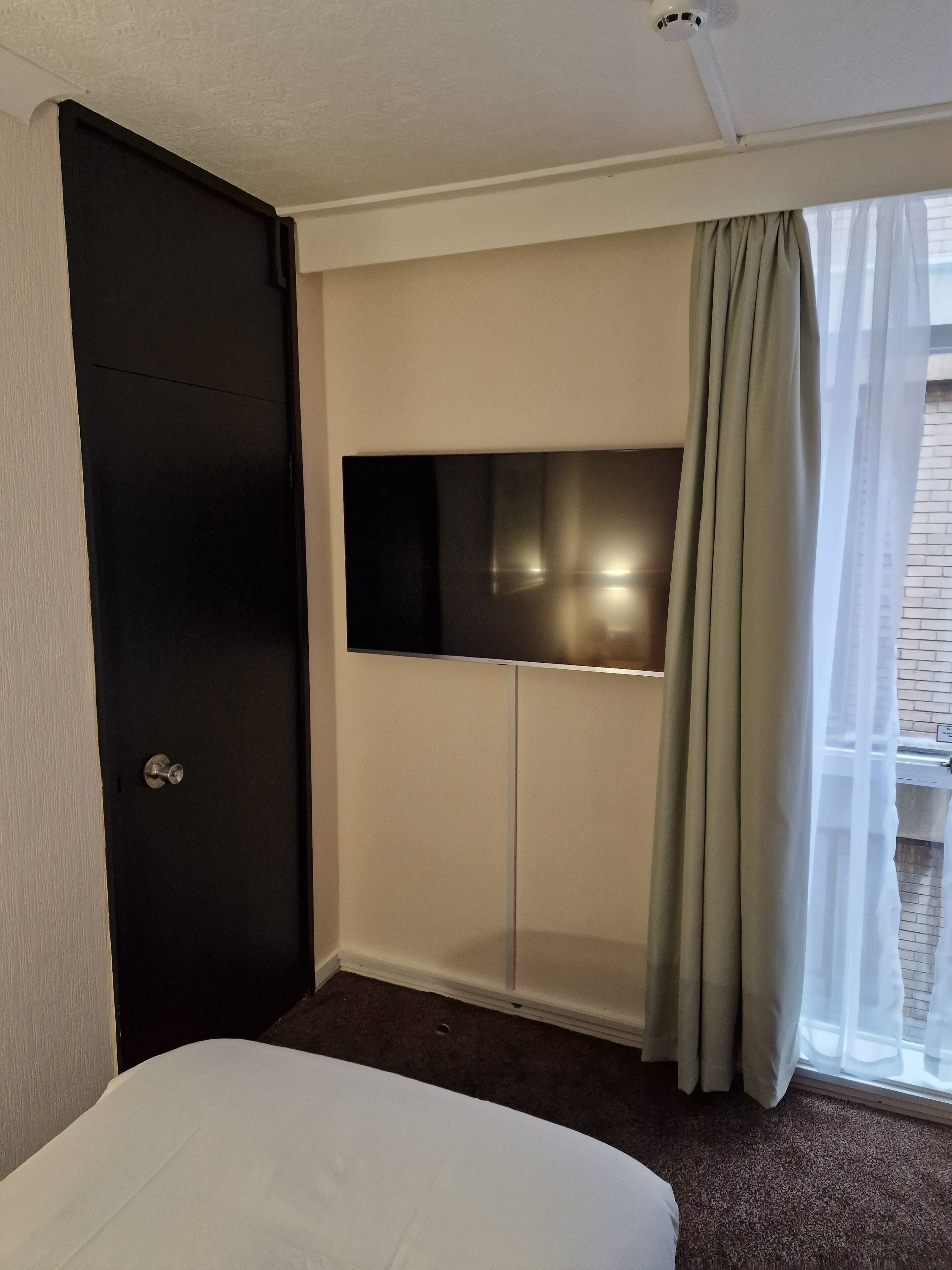 A hotel room with a bed, a TV mounted on a wall, a door, and a window with curtains.
