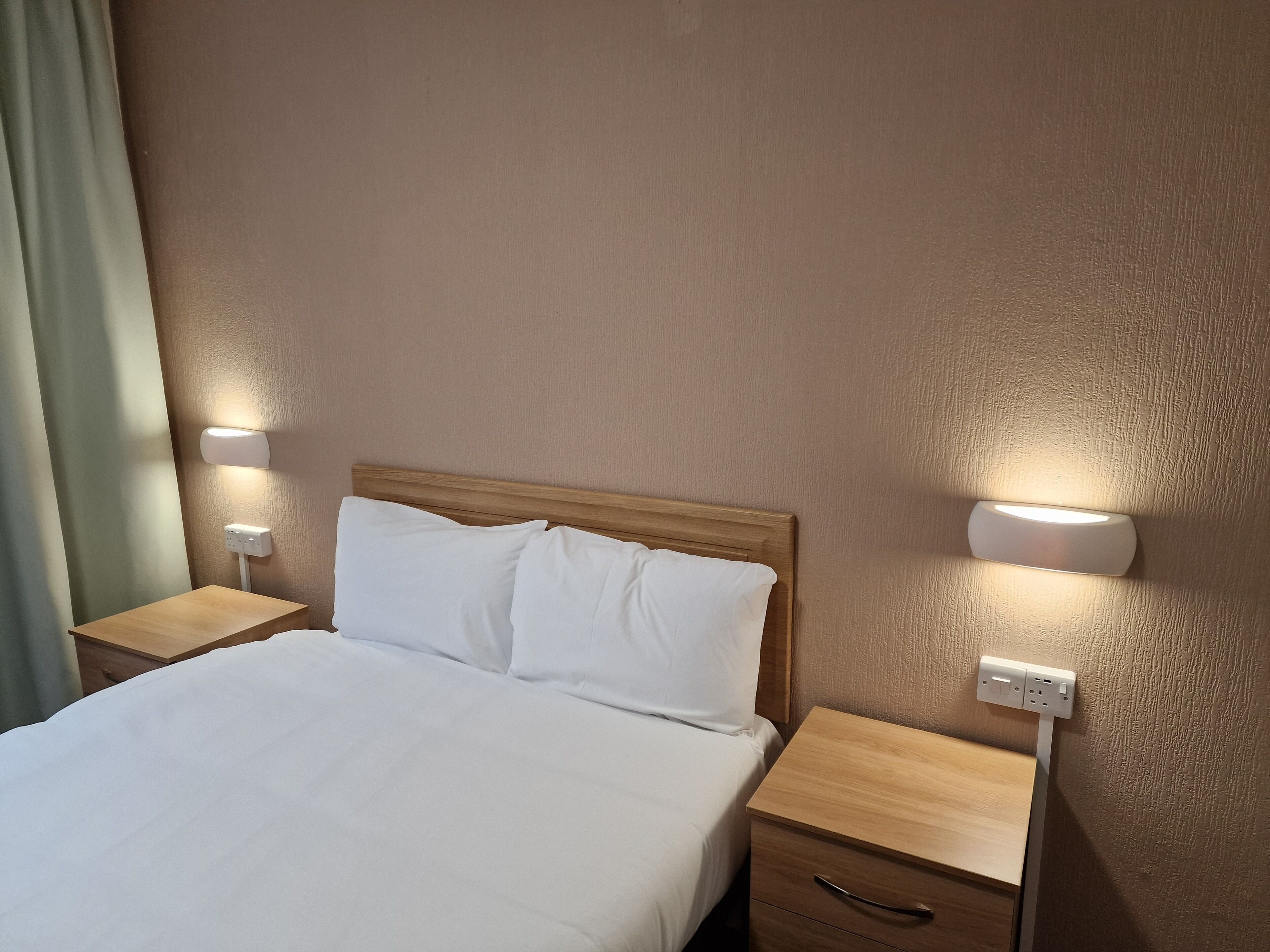 A hotel room with a wooden headboard, white bedding, two bedside tables with lamps, and a power outlet on the wall.