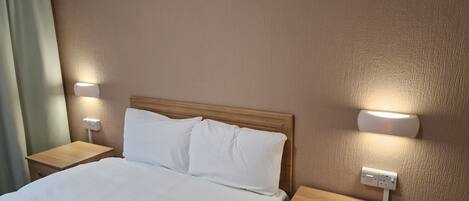 A hotel room with a wooden headboard, white bedding, two bedside tables with lamps, and a power outlet on the wall.