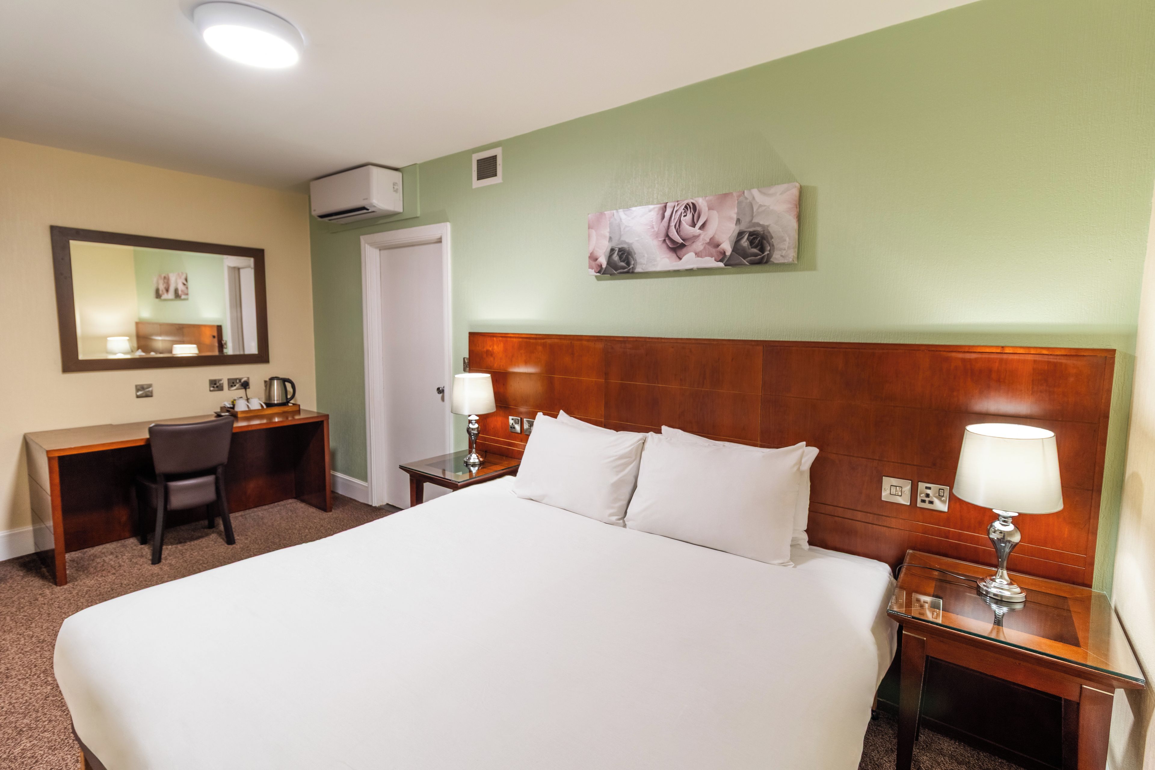 A hotel room with a large bed, a desk, a chair, a nightstand, and a wall-mounted air conditioning unit.