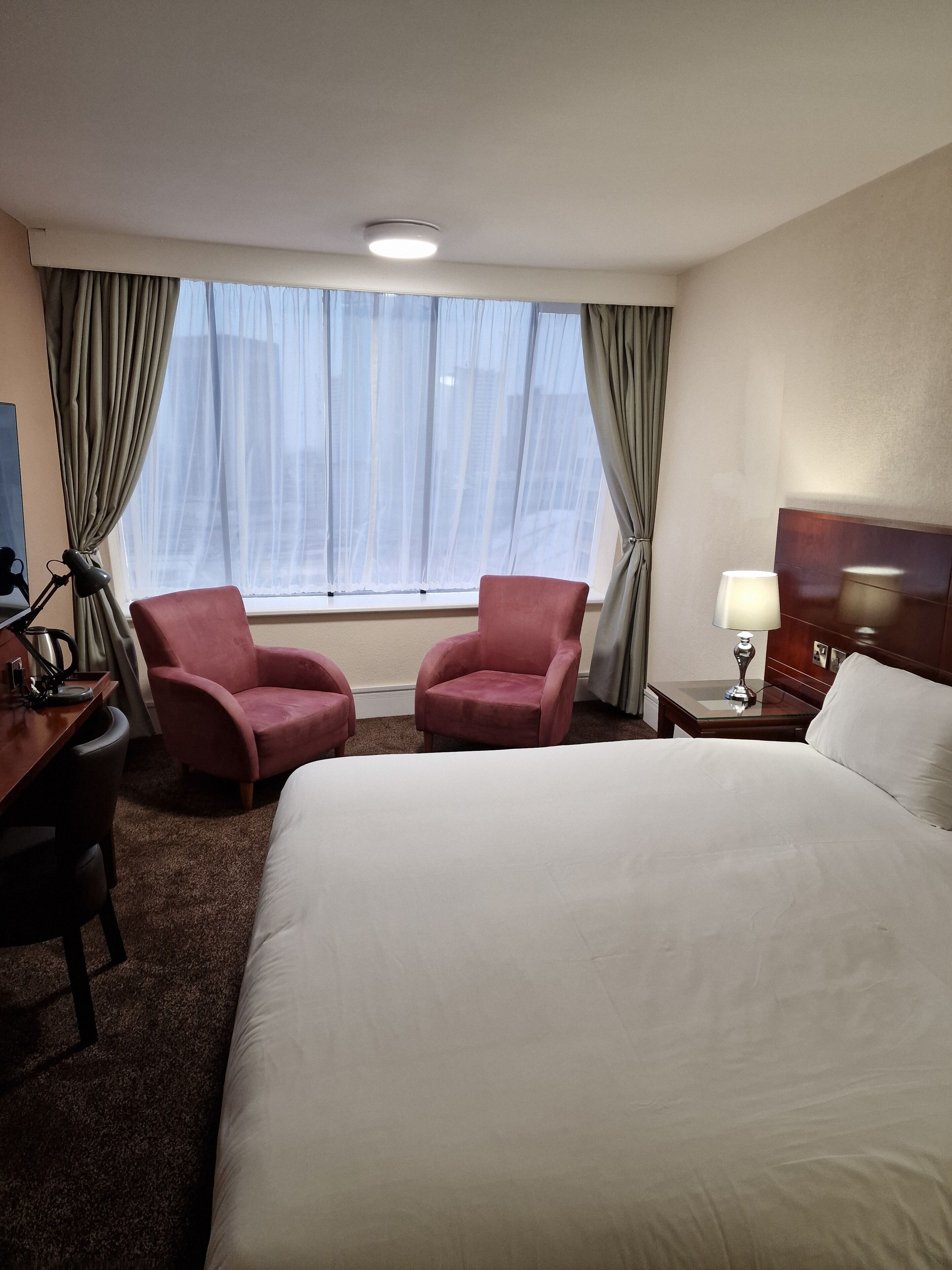 Et hotelværelse med en stor seng, to lænestole, et skrivebord med lampe og et sengebord med lampe.