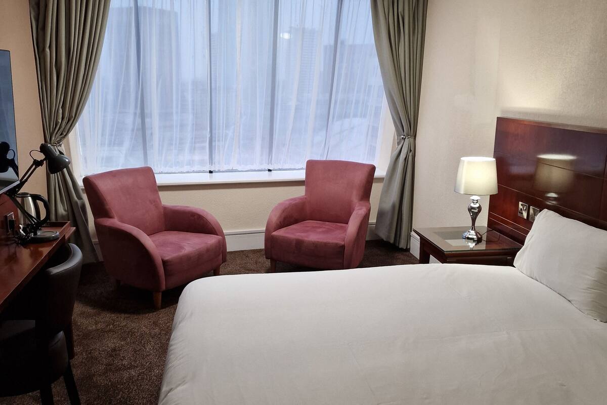 Una habitación de hotel con una cama grande, dos sillones, un escritorio con lámpara y una mesita de noche con lámpara.