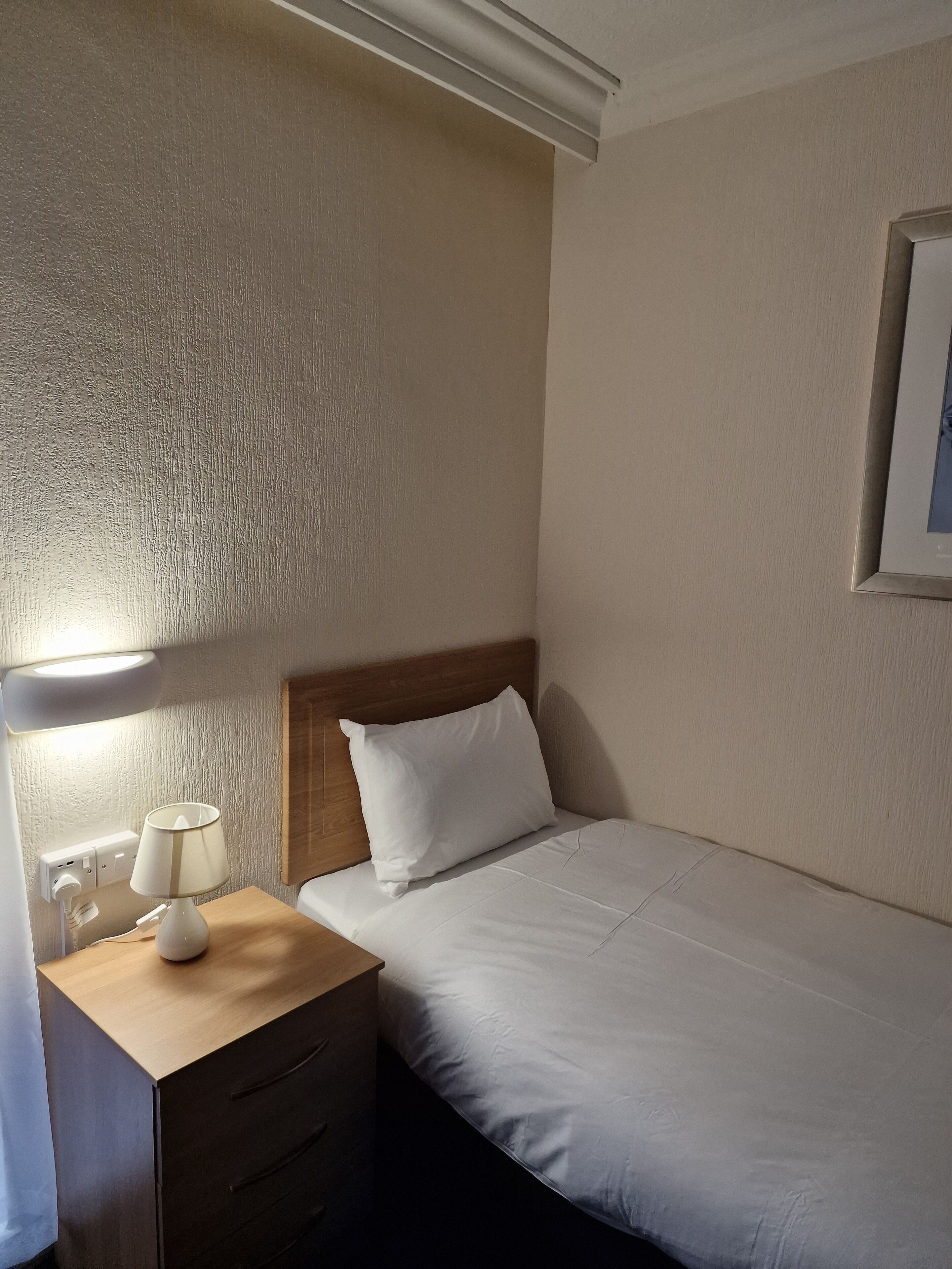 A hotel room with a wooden nightstand, a white lamp, a single bed with white bedding, and a framed picture on the wall.
