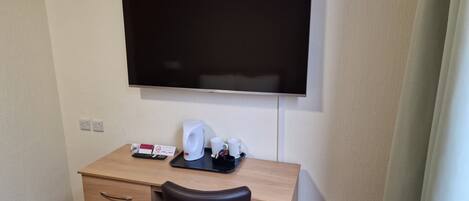 A hotel room with a wooden desk, a black leather chair, a flat-screen TV mounted on the wall, a coffee maker, and a remote control.