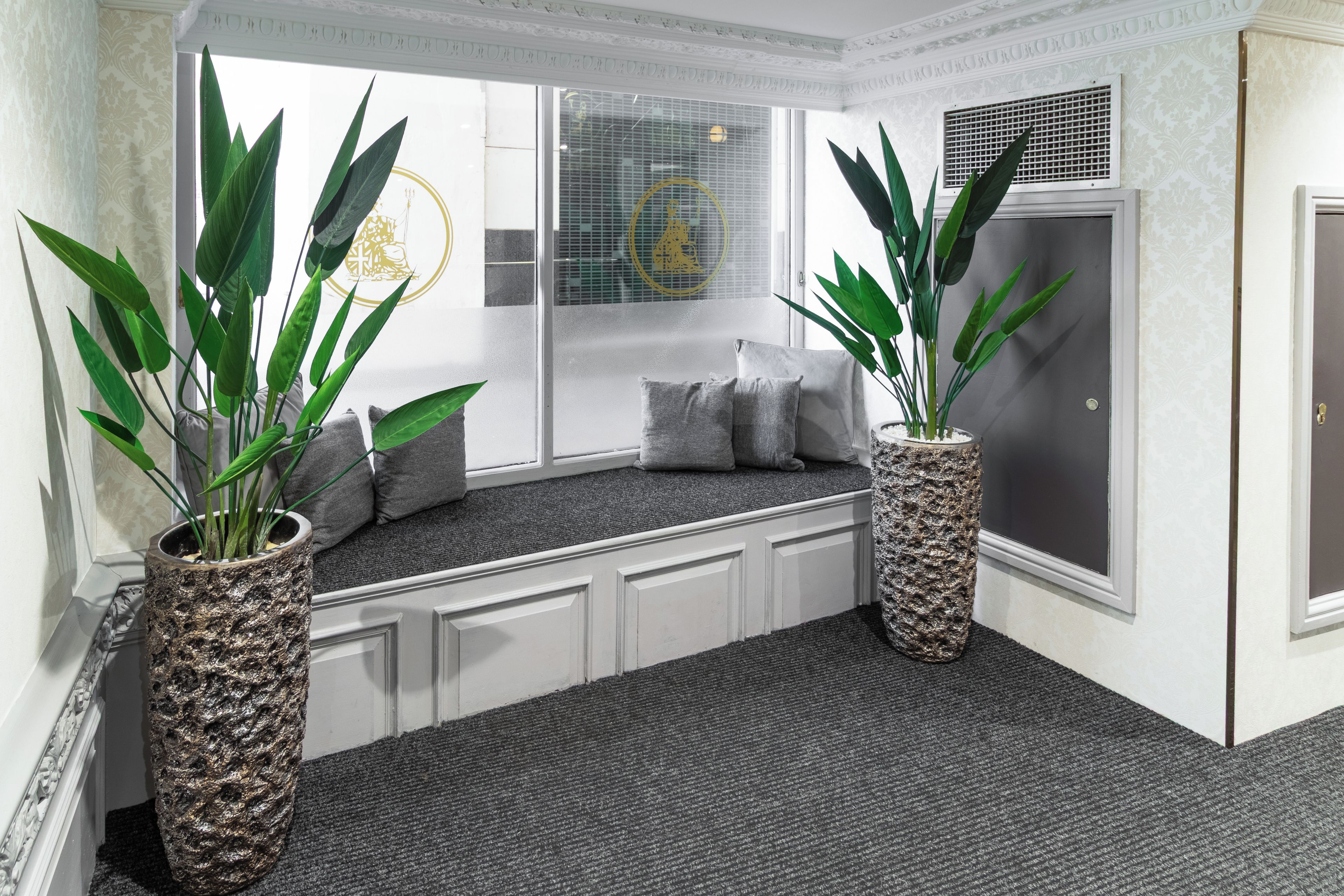A room with a built-in bench, two large potted plants, a window with blinds, and a door with a textured wall.
