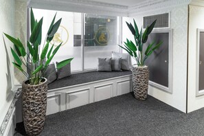 Una habitación con un banco integrado, dos plantas grandes en macetas, una ventana con persianas y una puerta con pared texturizada.