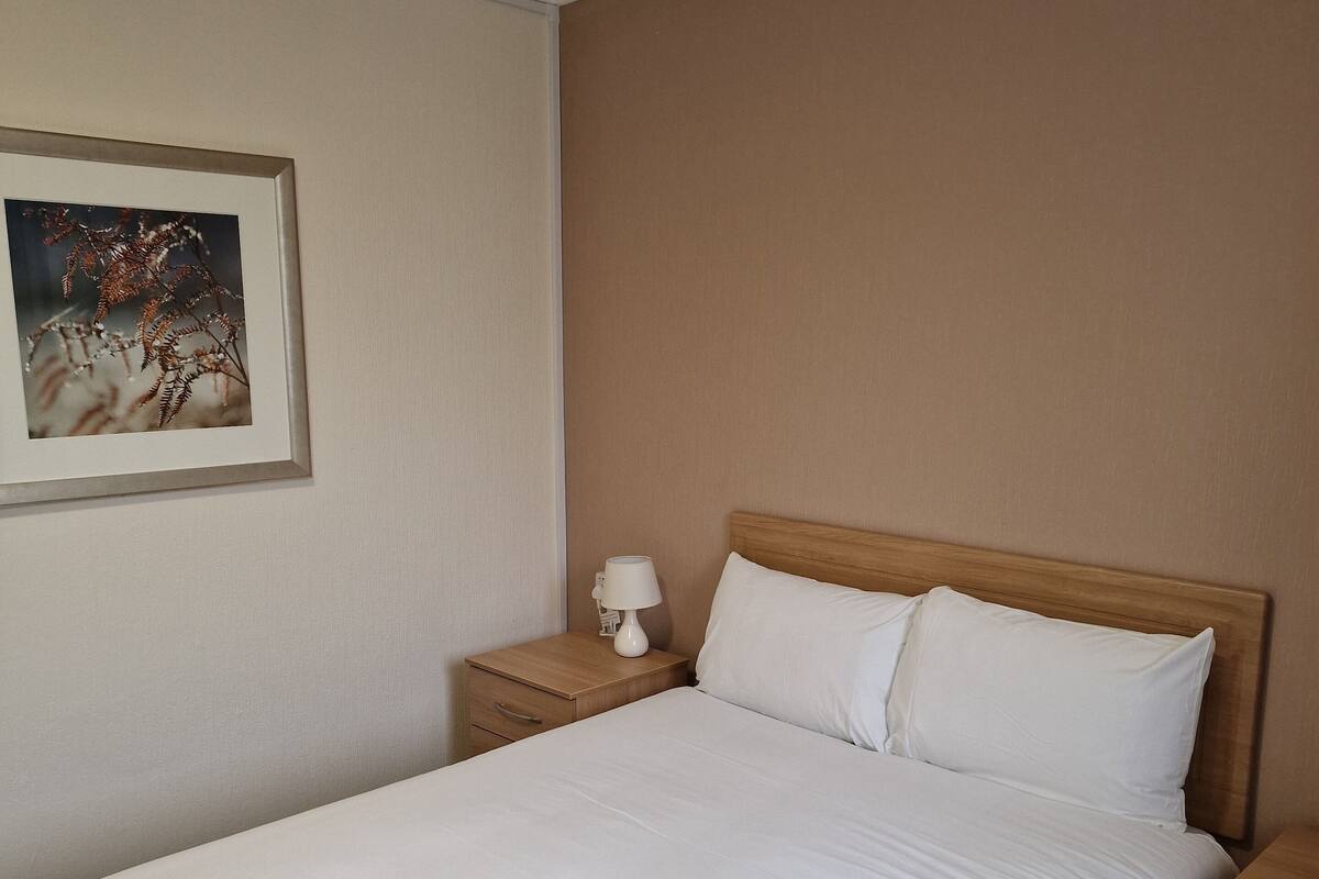 Una cama bien hecha con sábanas blancas, cabecera de madera y mesita de noche con lámpara.