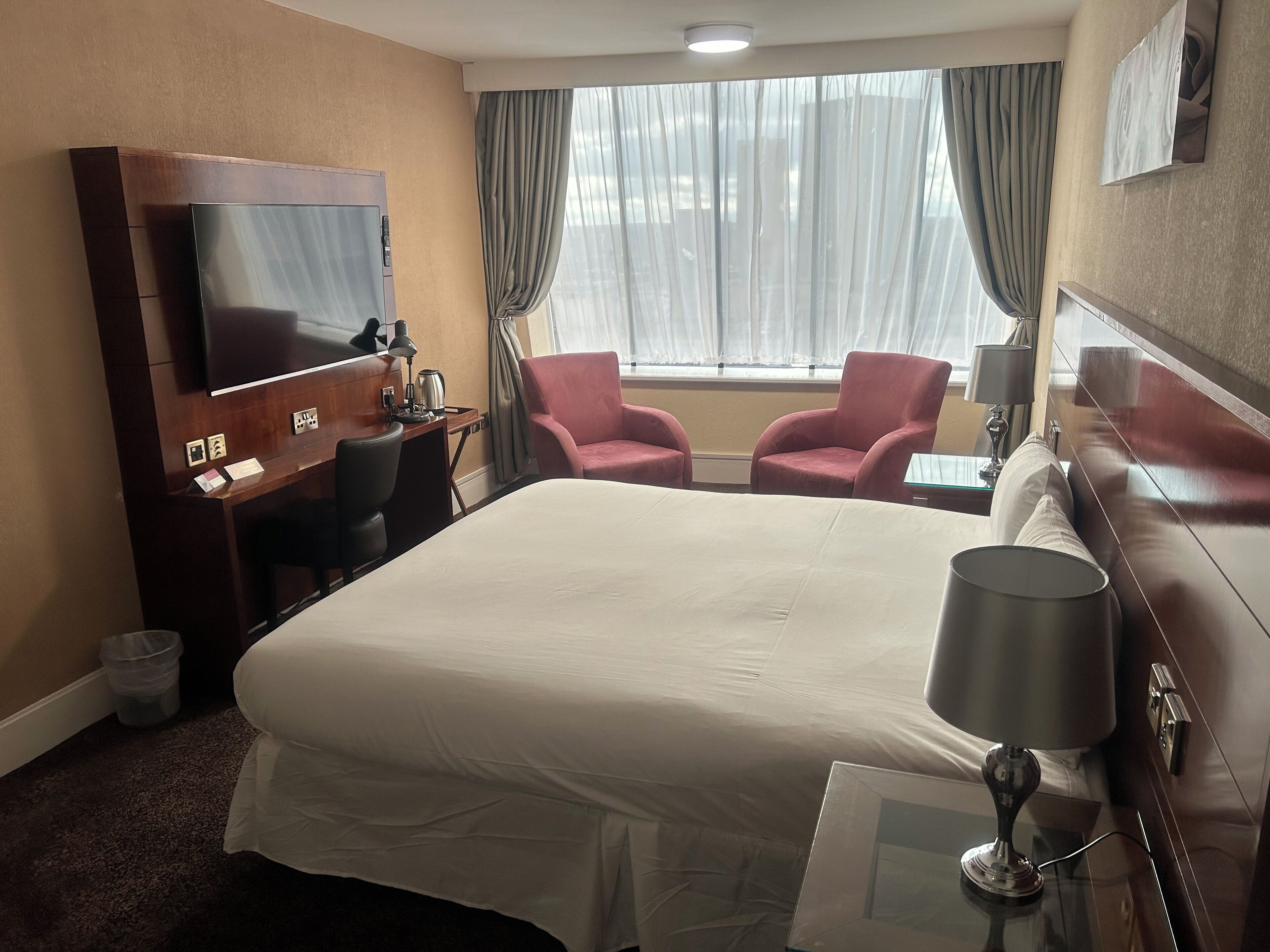 A hotel room with a large bed, a desk with a flat-screen TV, two pink armchairs, and a window with sheer curtains.