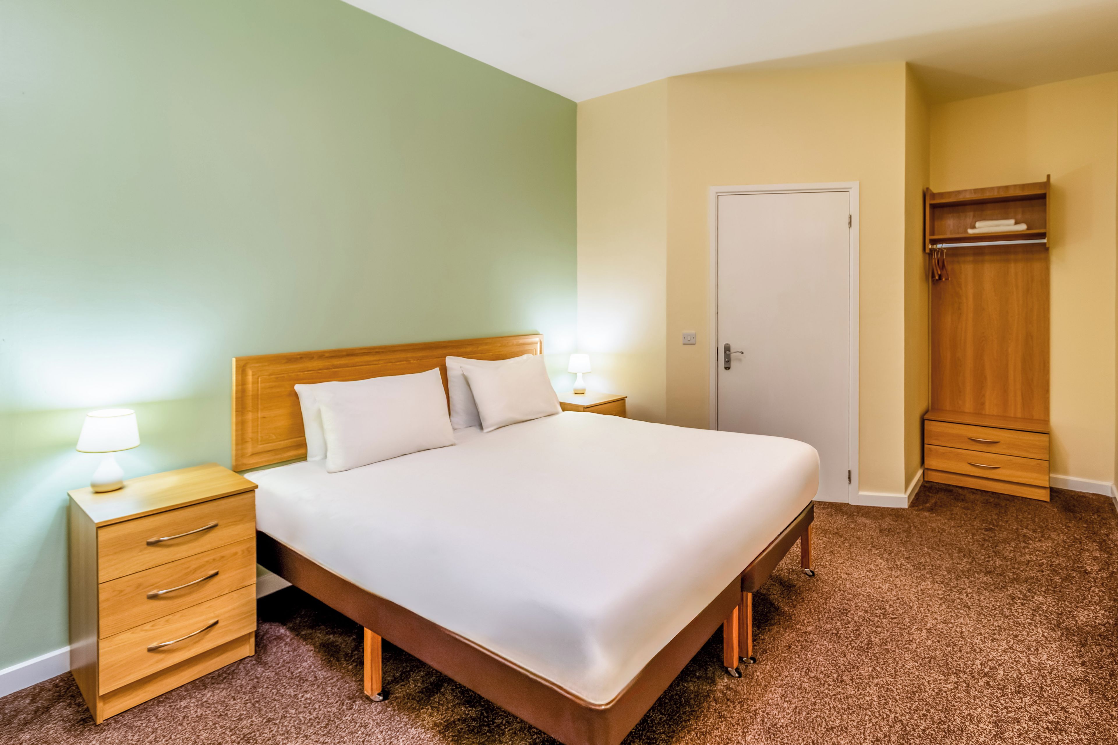 A neatly made bed with a wooden headboard, two bedside tables with lamps, a wardrobe, and a door.