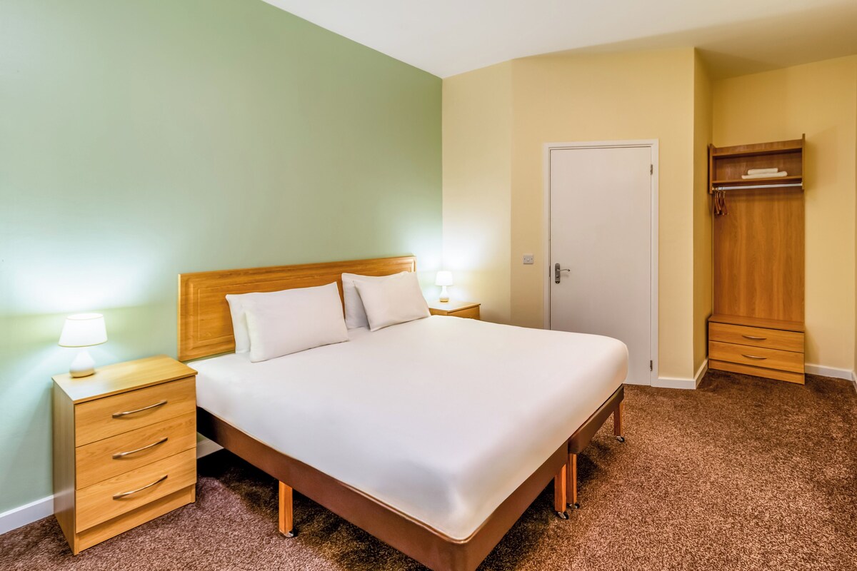 Una cama bien hecha con cabecera de madera, dos mesitas de noche con lámparas, un armario y una puerta.