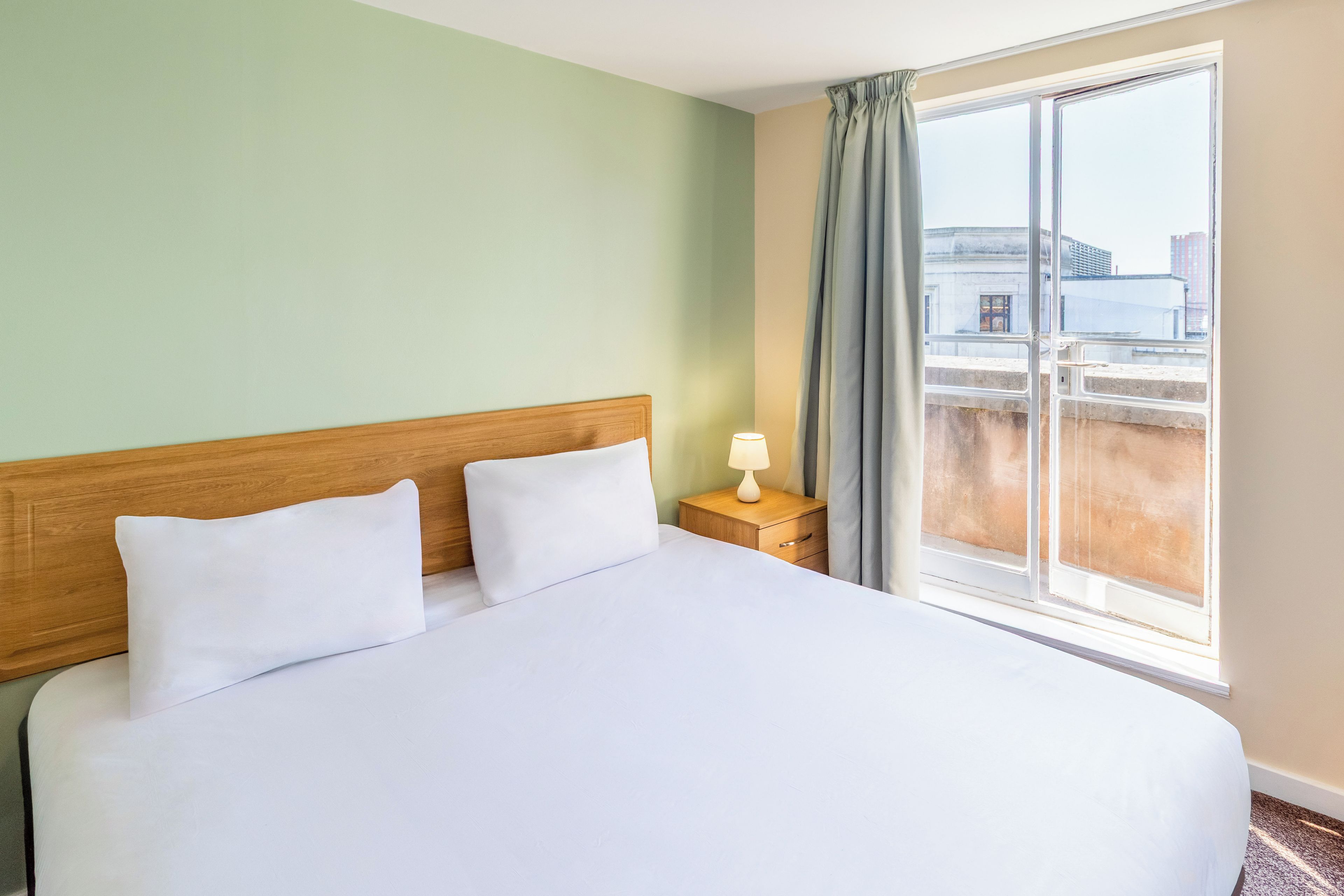 A hotel room with a bed, a nightstand, a lamp, and a window with a view of buildings.