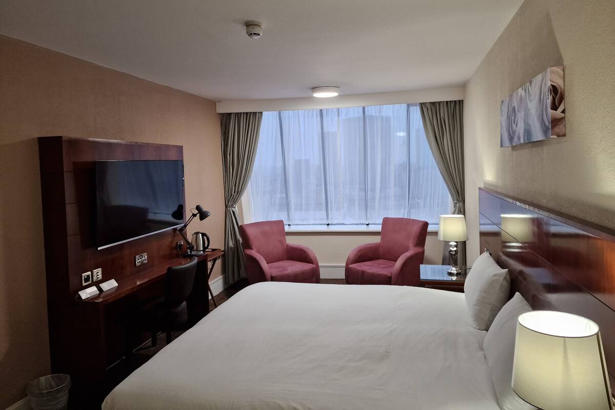 Habitación de hotel con cama, dos sillones, televisor, escritorio con lámpara y ventana con cortinas.