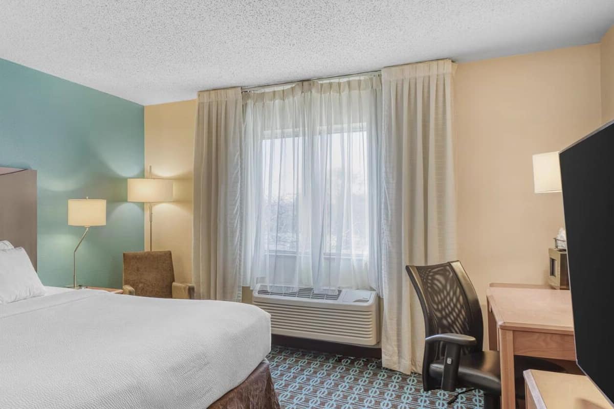 A hotel room with a bed, a desk, a chair, a lamp, and a window with curtains.