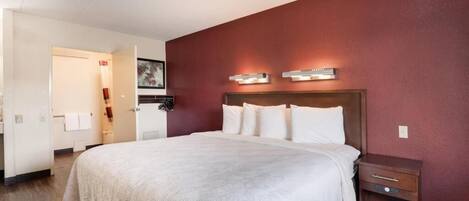 Quarto de hotel com uma cama grande, uma mesinha de cabeceira de madeira, uma luminária fixa na parede e uma porta que dá para um banheiro.