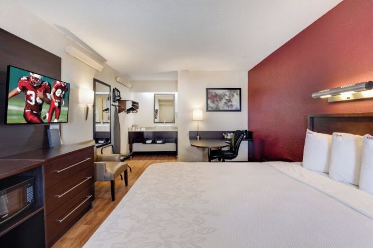 A hotel room with a large bed, a desk with a chair, a television, a mirror, and a red accent wall.