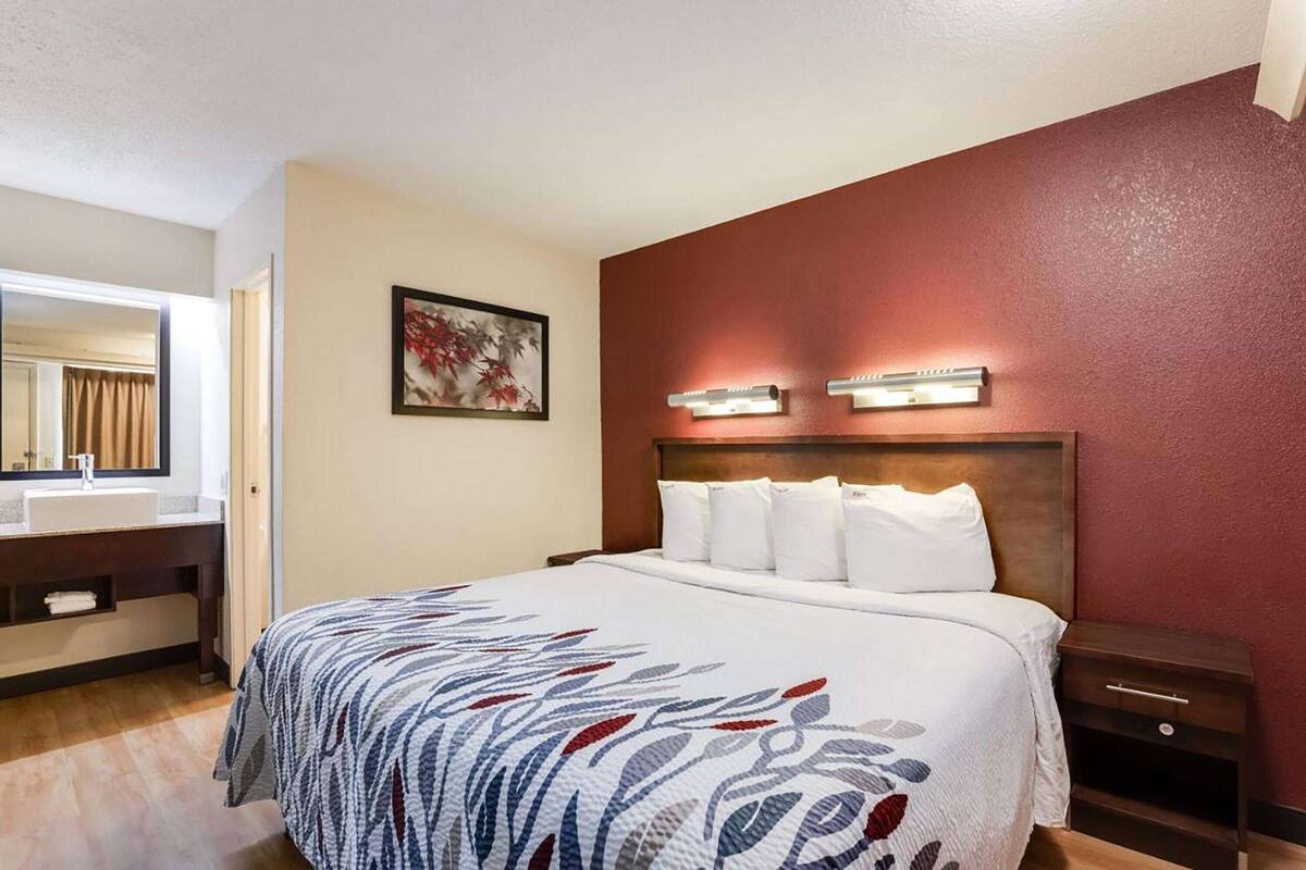 Une chambre d’hôtel comprenant un lit, une table de chevet, une commode, une salle de bain avec un lavabo et un miroir, et un luminaire fixé au mur.