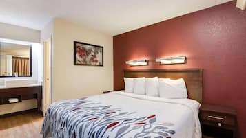 Una habitación de hotel con cama, mesita de noche, lavabo y espejo en el baño, y una lámpara fijada a la pared.