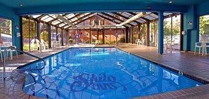 Una piscina interior con agua cristalina, rodeada por una zona techada con sillas y mesas.