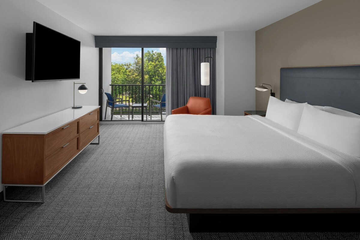 A hotel room with a large bed, a TV mounted on the wall, a desk with drawers, a chair, and a balcony with a view of trees.