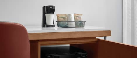 A desk with a coffee maker, a trash bin, and a mesh basket containing two packets labeled "Rainforest Blend."