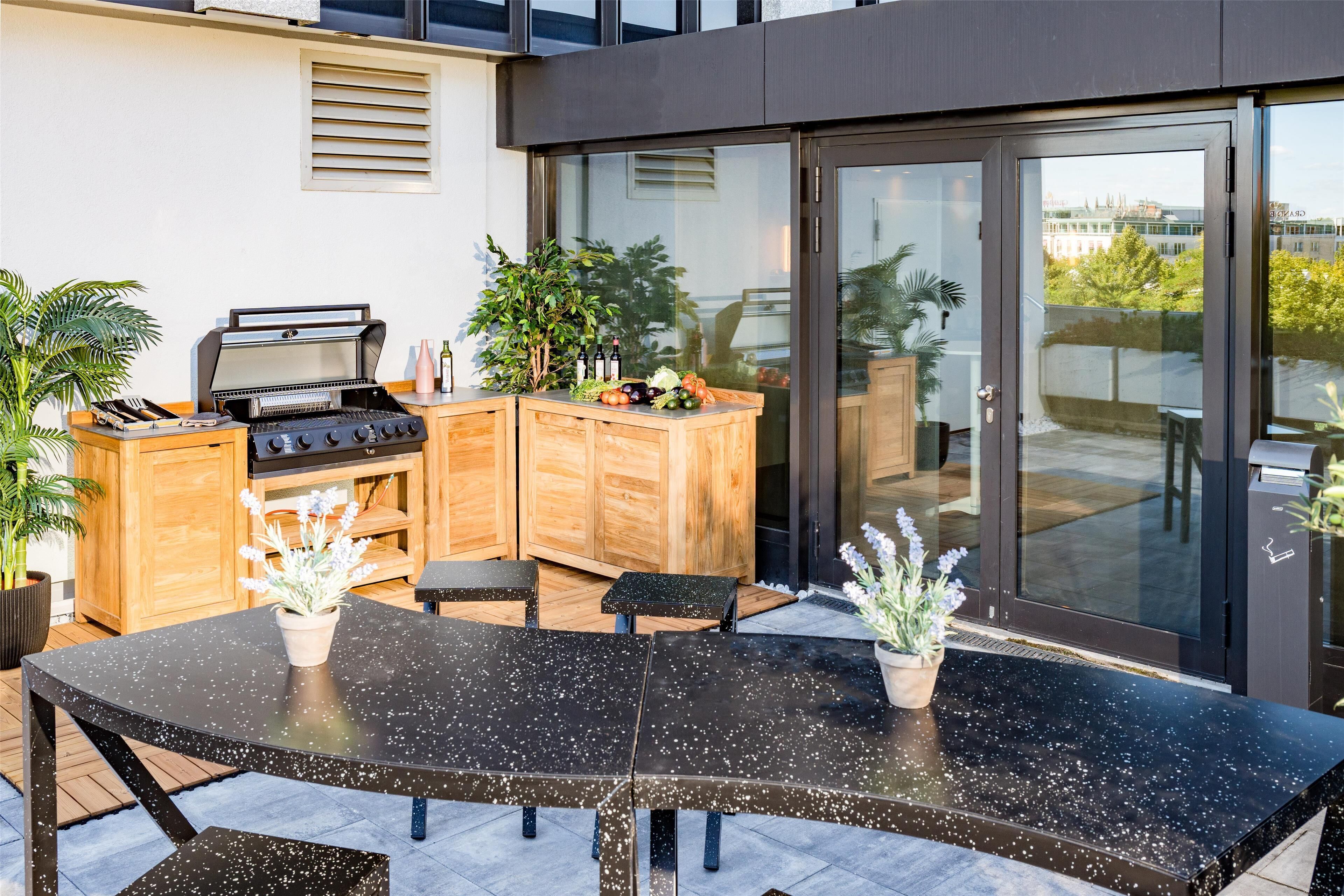 Een moderne buitenkeuken met een barbecue, houten kastjes en een eethoek met een tafel en stoelen met een zwart gespikkelde afwerking.
