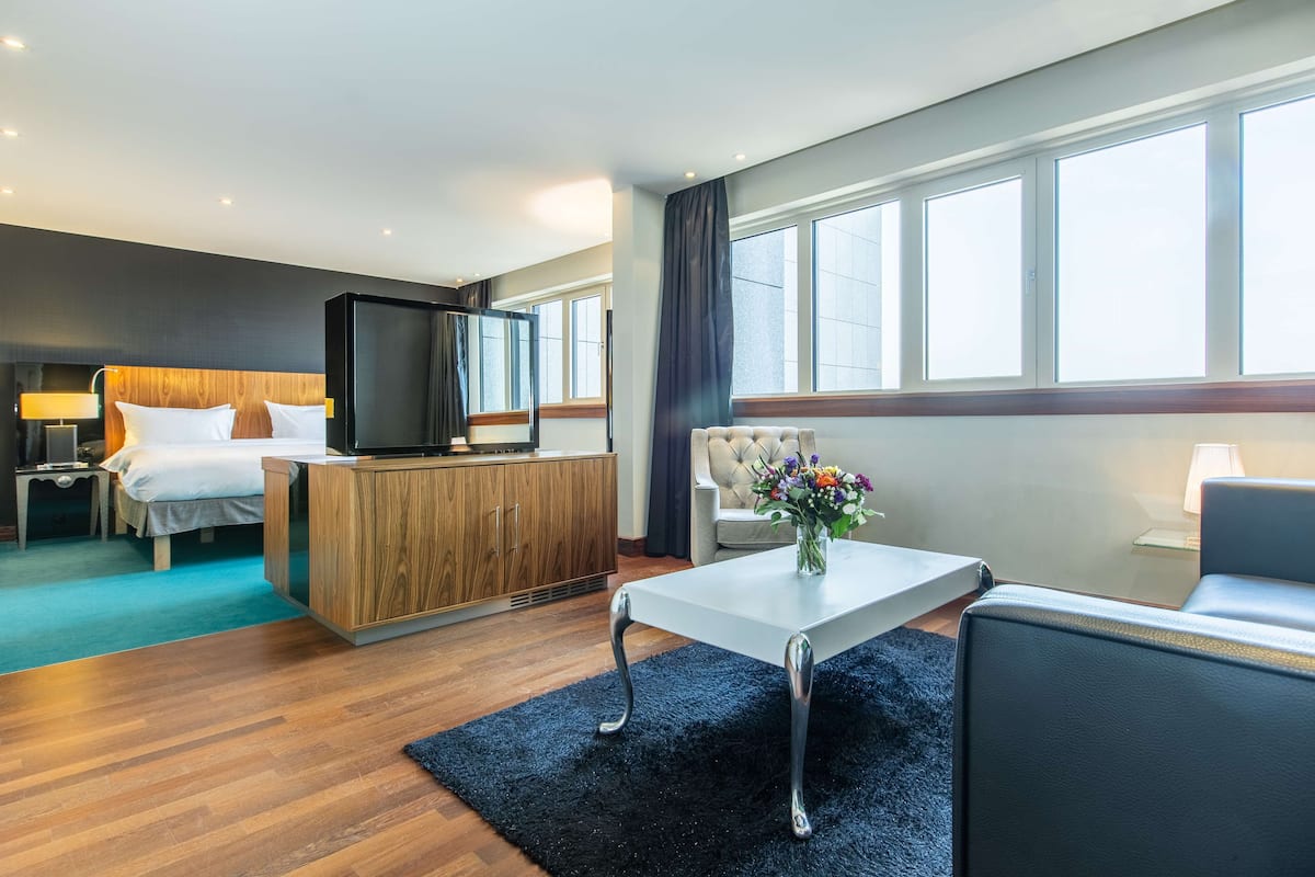 A modern hotel room with a bed, a TV, a wooden cabinet, a sofa, a coffee table, and large windows.