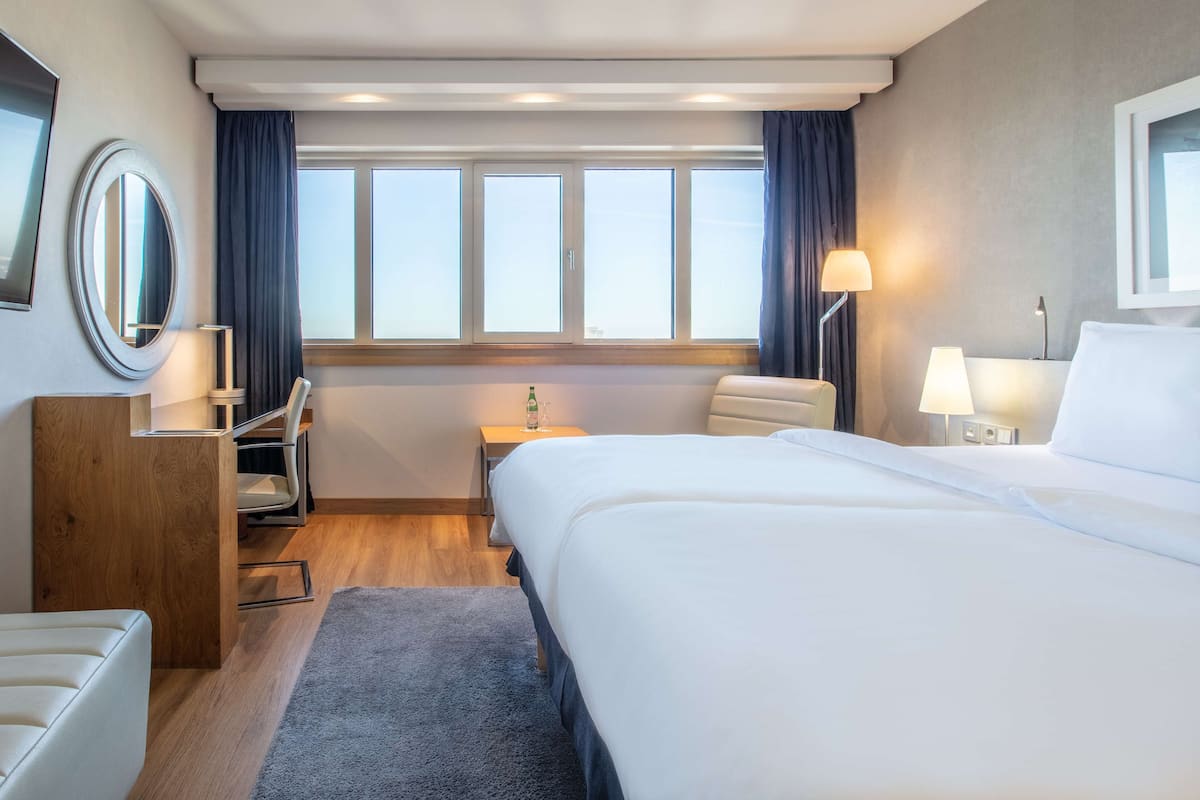 A hotel room with a large bed, a desk with a chair, a small table with a lamp, and a window with curtains.