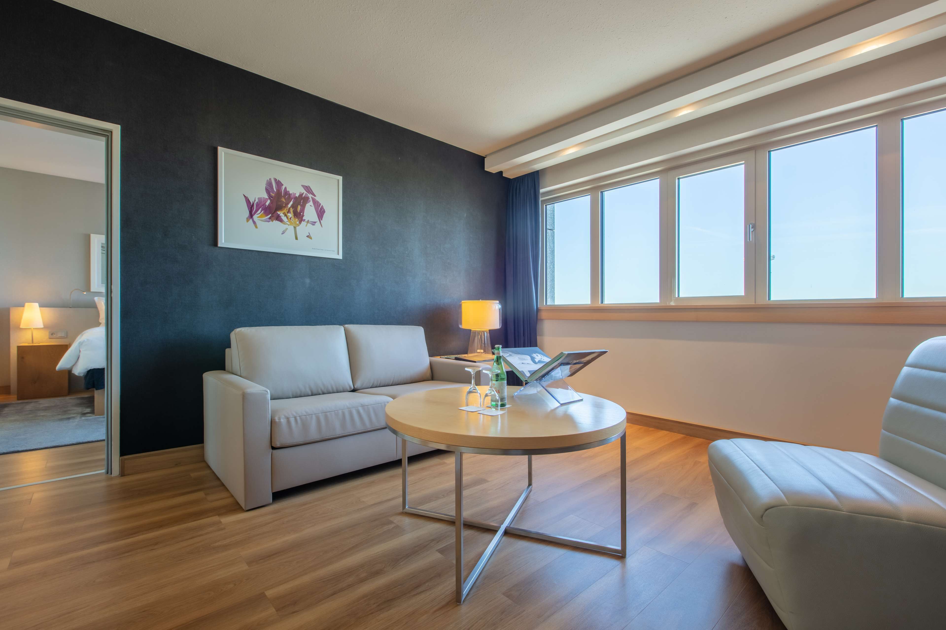 Moderni hotellihuone, jossa on sohva, pyöreä pöytä lampun kanssa ja suuri ikkuna, jossa on verhot.