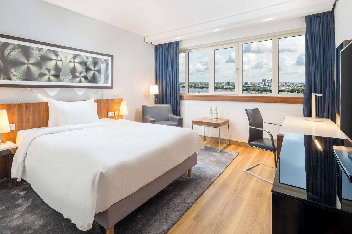 A modern hotel room with a large bed, a desk, a chair, a TV, and a window with a city view.