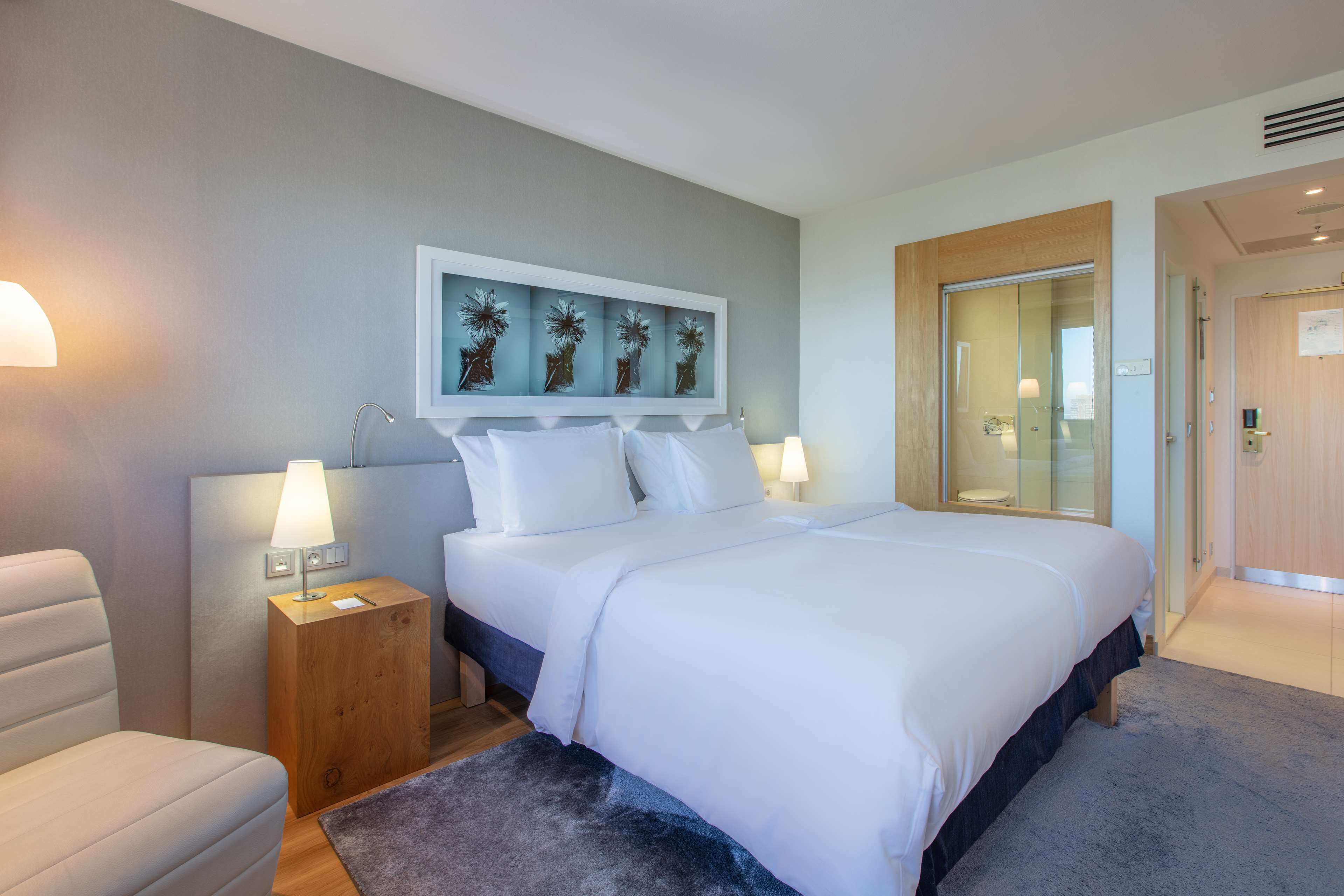 Una habitación de hotel moderna con una cama grande, una mesita de noche de madera, una lámpara de escritorio y una obra de arte en la pared.