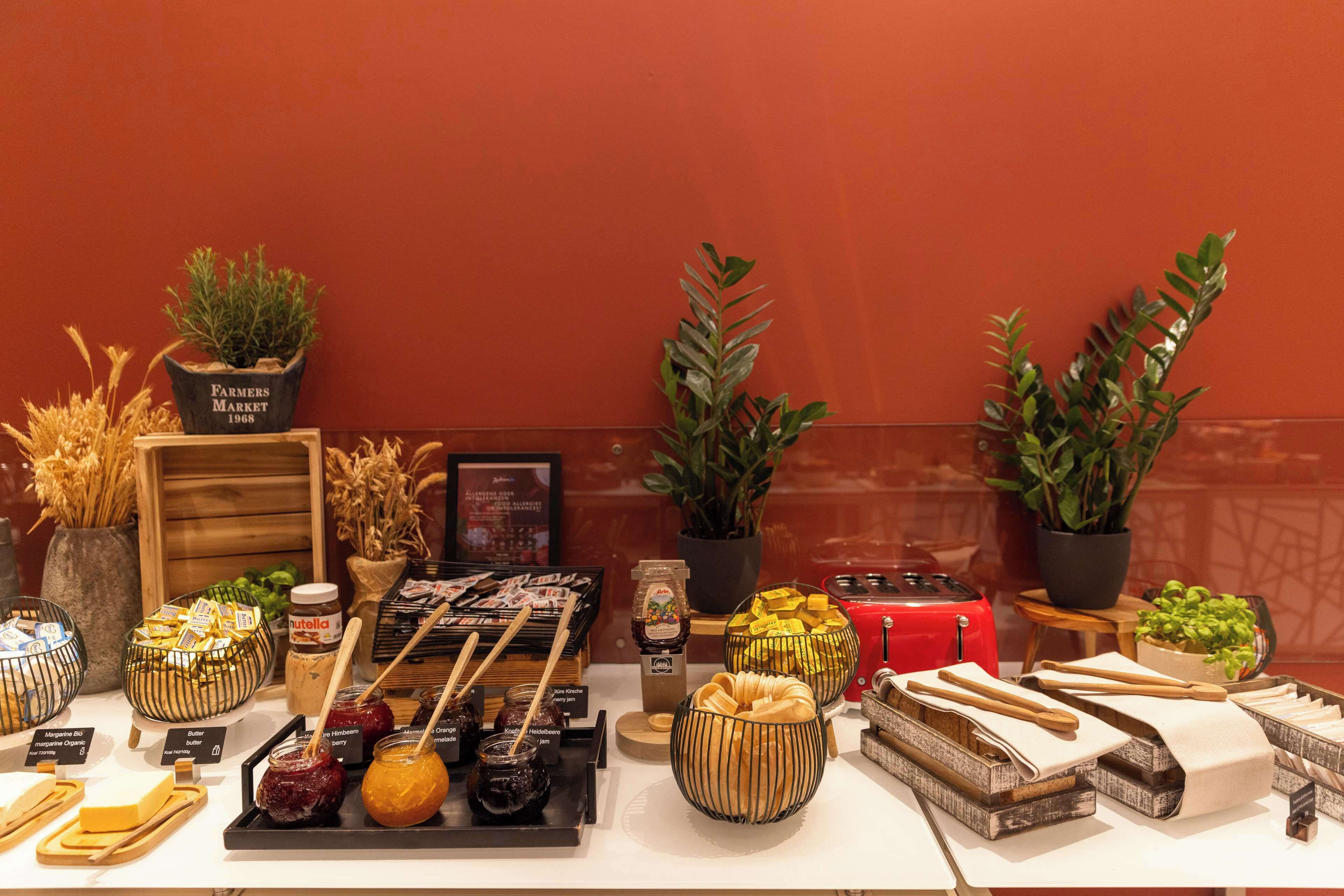 A buffet table with various food items, including cheeses, jams, and pastries, arranged on a wooden table with potted plants and a red toaster.