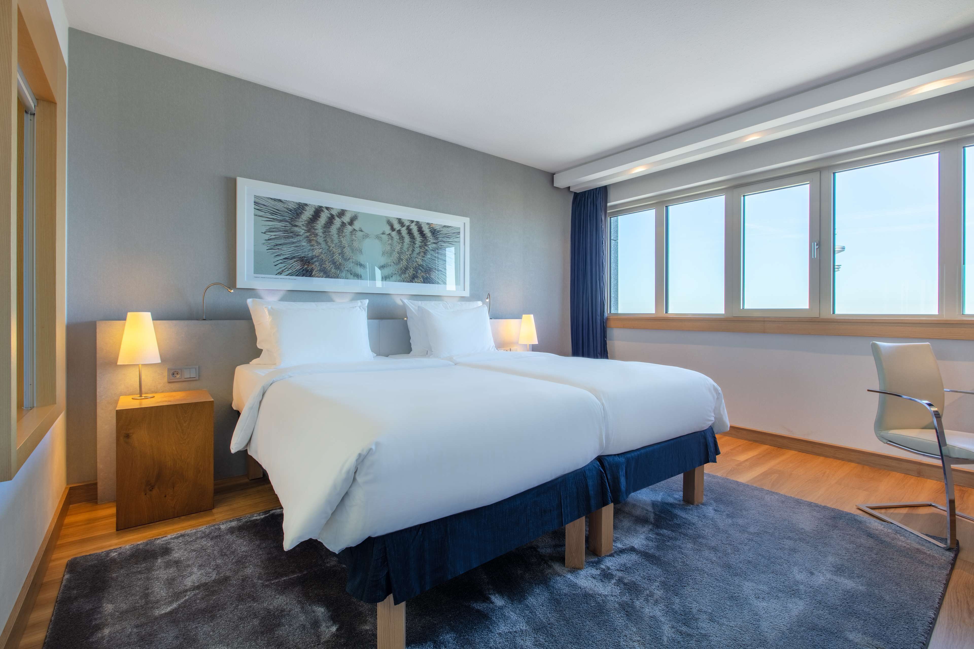 A modern hotel room with a large bed, two bedside tables with lamps, a chair, a large window, and a framed artwork on the wall.