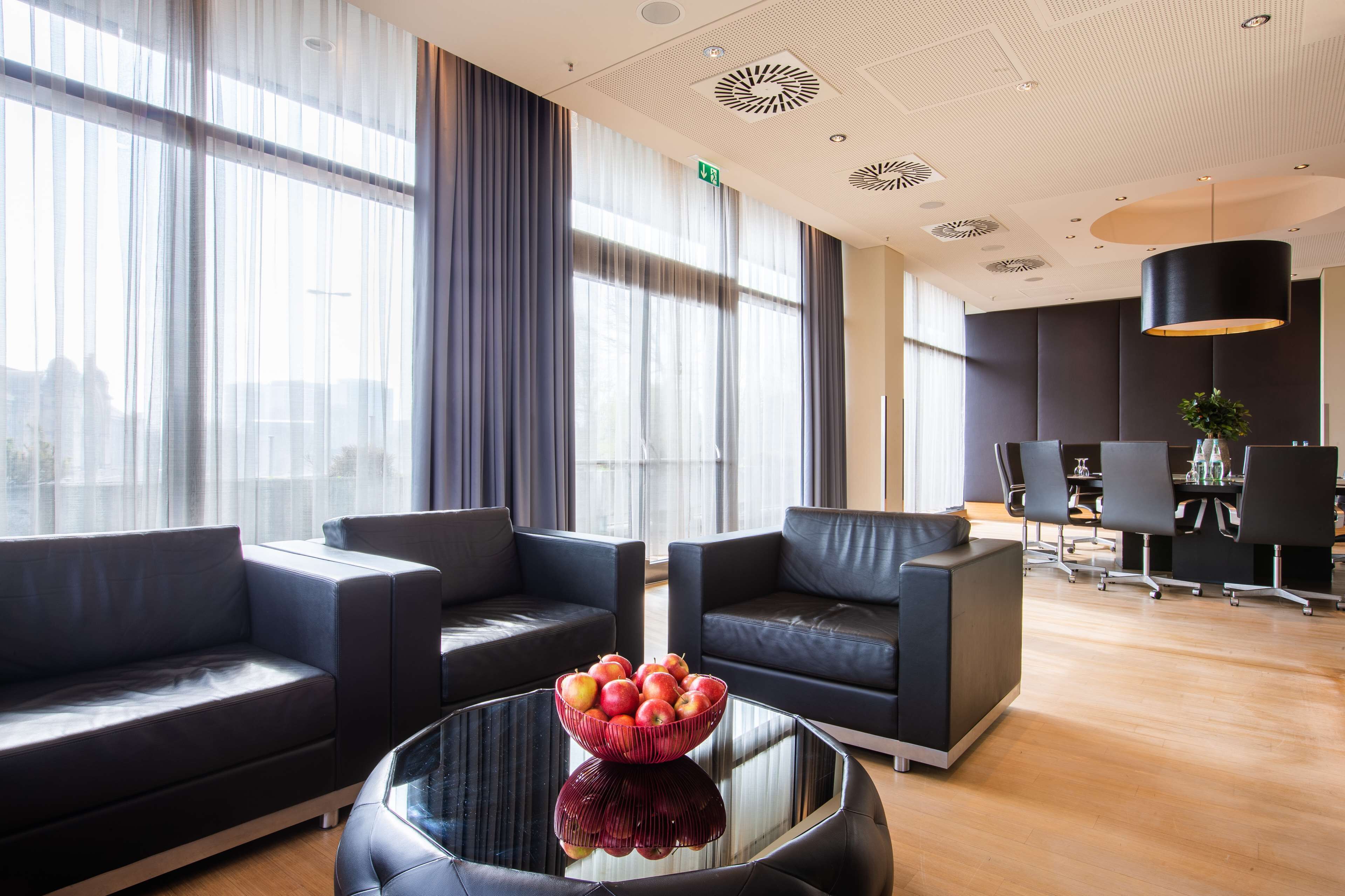 Un hall d’entrée moderne avec des canapés en cuir, une table basse en verre ornée de fruits, de grandes fenêtres et un espace dédié aux réunions.