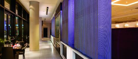 A modern hotel corridor with a long table set for dining, a large illuminated wall, and a clean, polished floor.