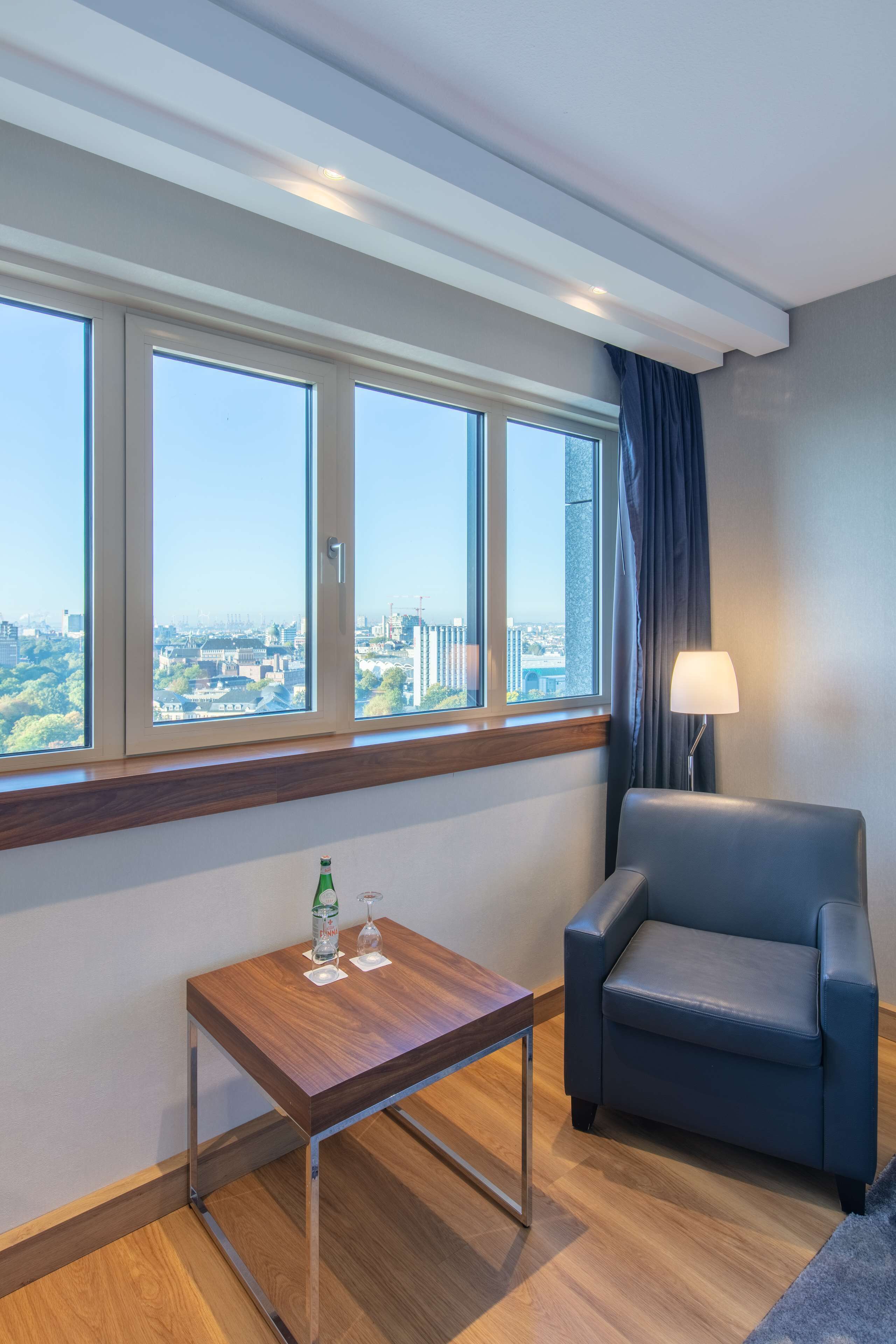 Habitación de hotel con un ventanal grande, una mesa de madera con una botella y un vaso, un sillón azul y una lámpara.